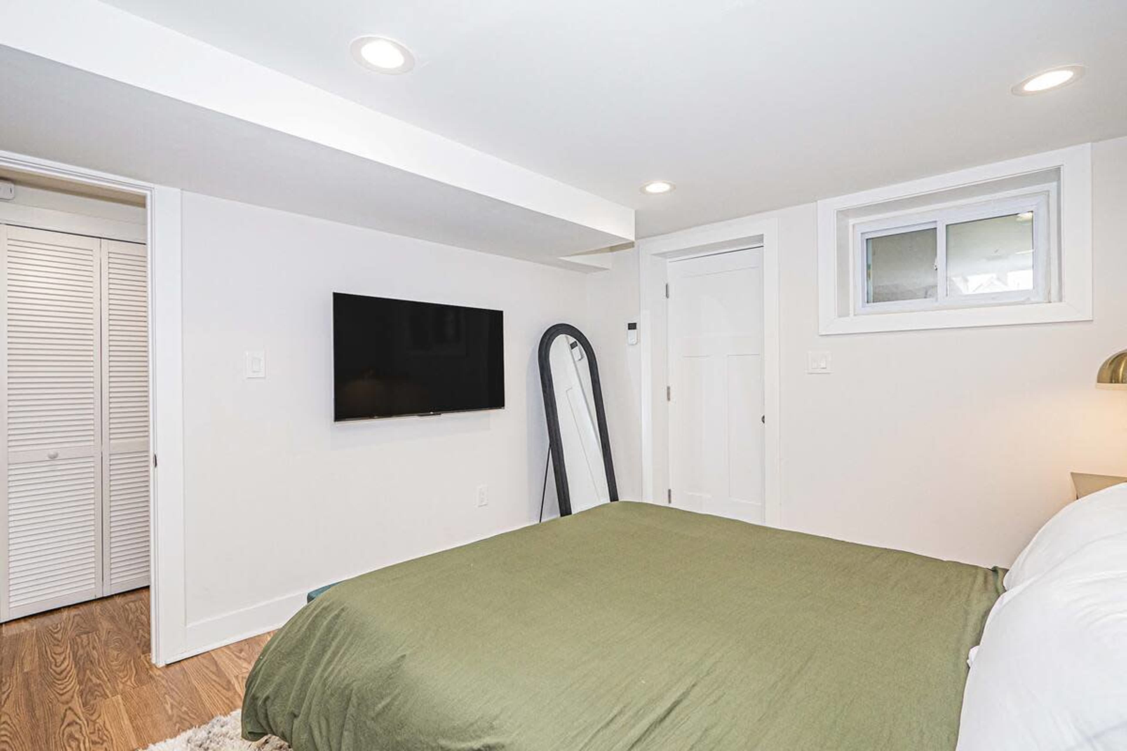 The image shows a modern, minimalist bedroom with a bed, a television mounted on the wall, and a door leading to a closet.