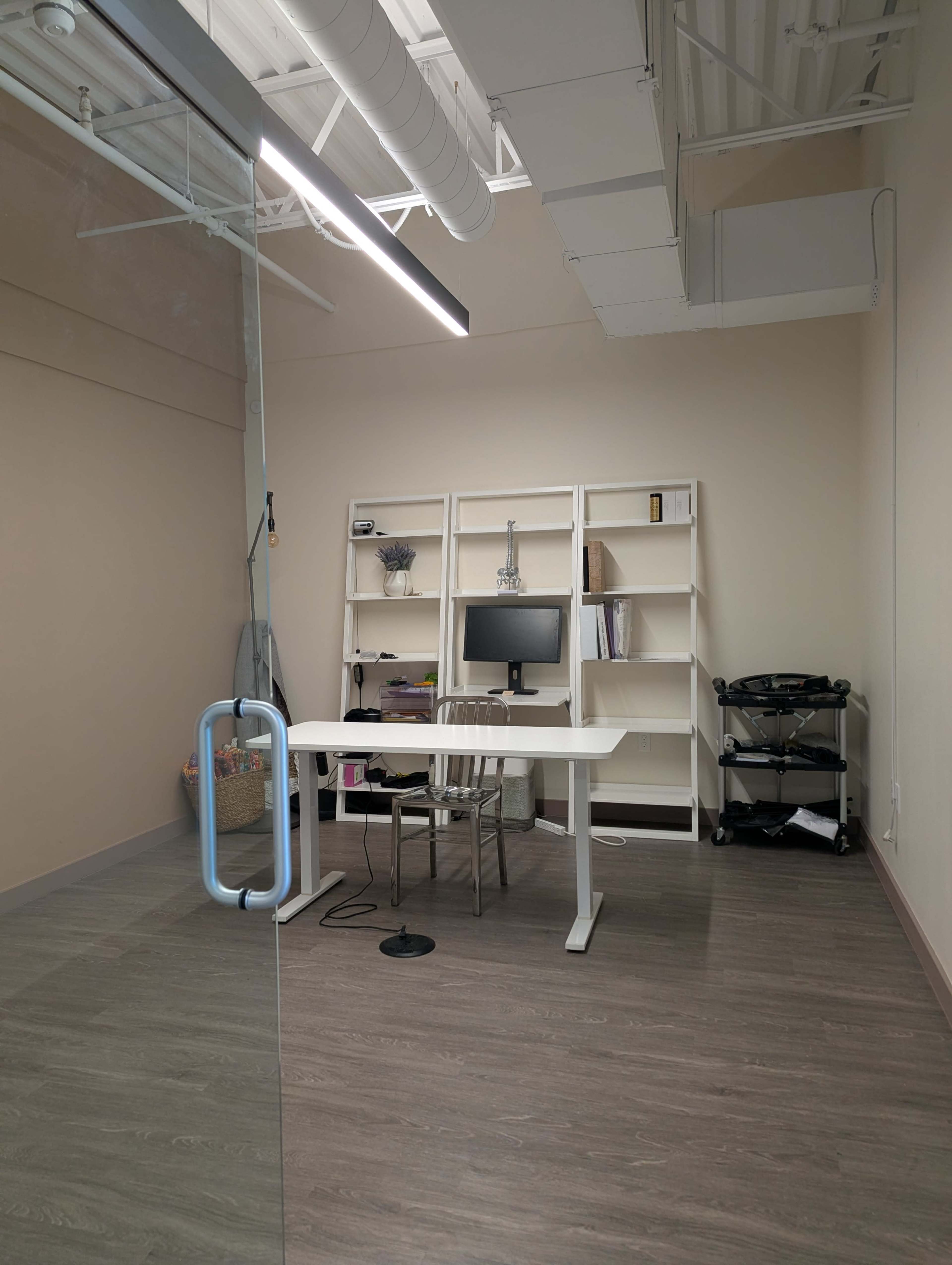 The image shows a minimalist office space featuring a desk and chair, a computer, shelving units, and a small cart, all set against light-colored walls.