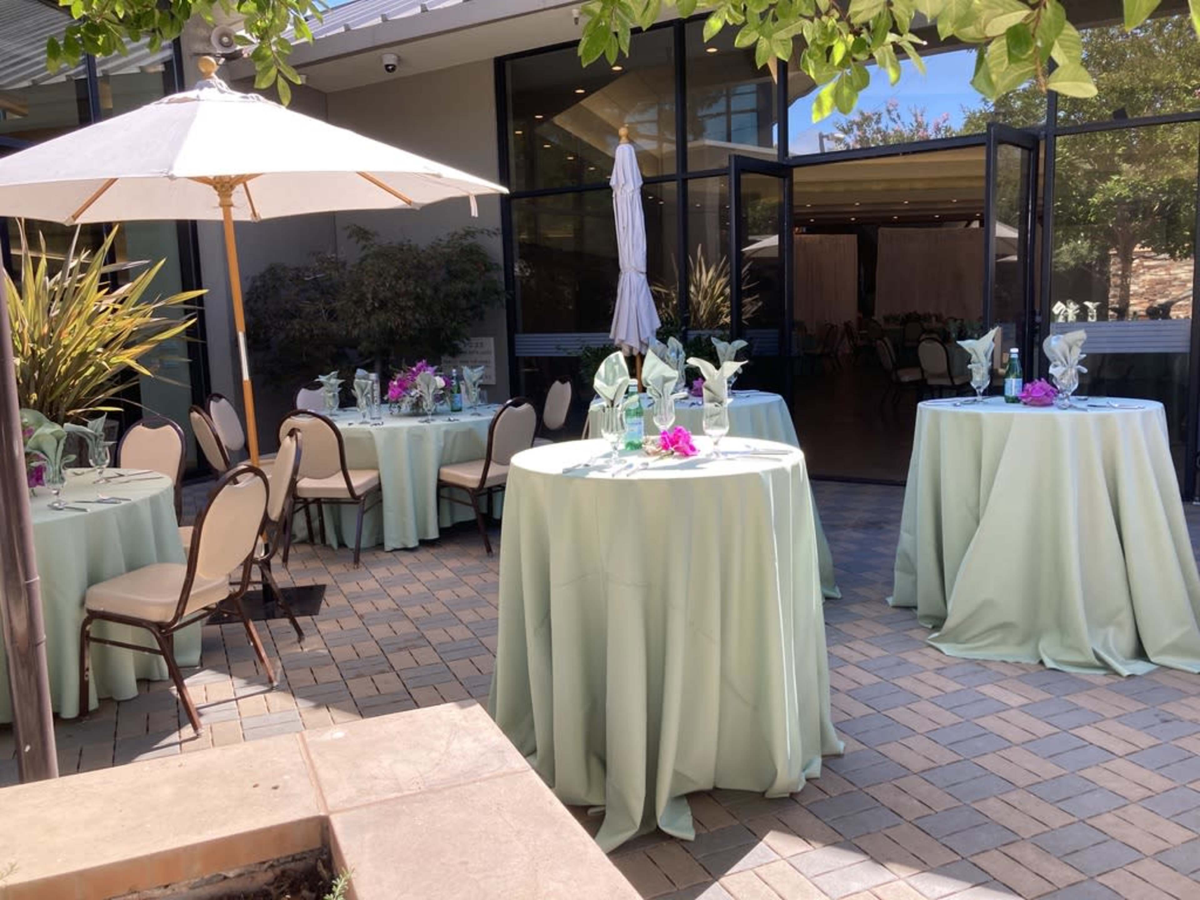 The image shows an outdoor seating area with several round tables covered in light green cloths, arranged with floral centerpieces and surrounded by elegant chairs.