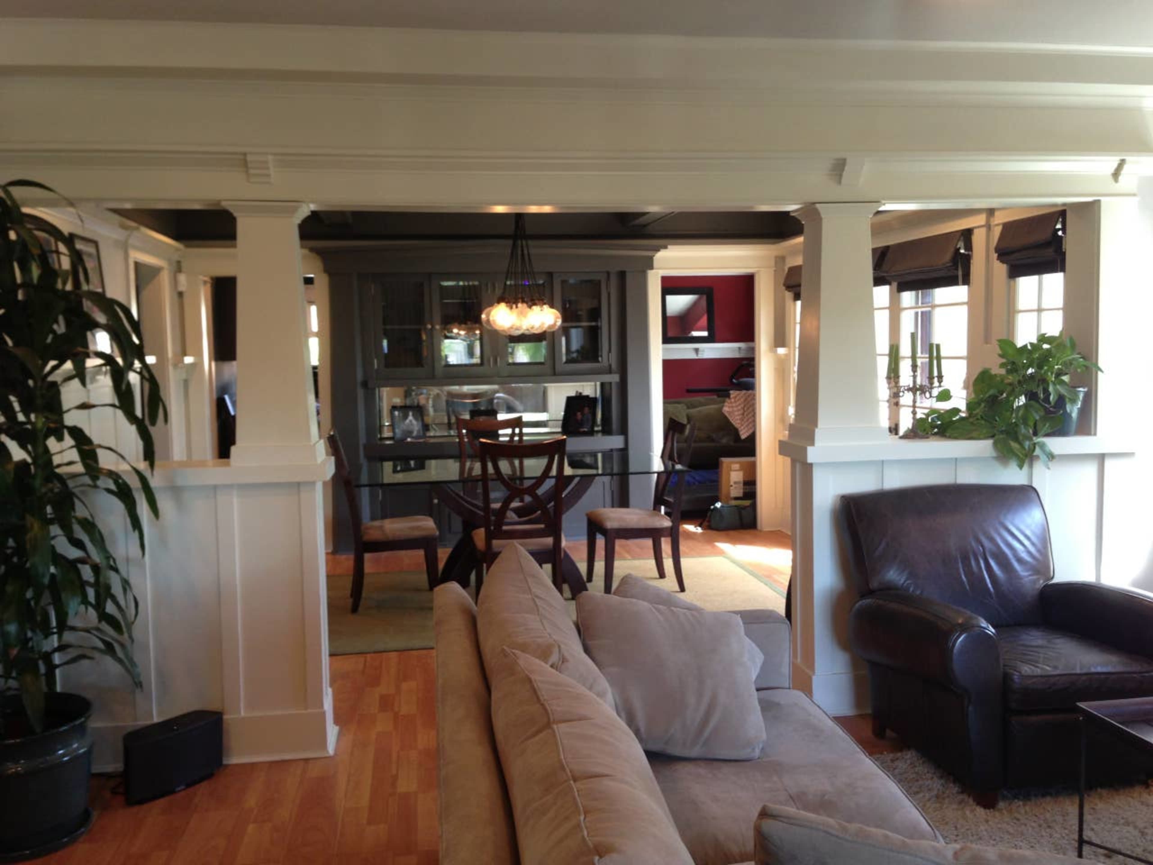 A spacious living area connected to a dining space, separated by white columns, with a large sofa, dark leather chairs, and several plants.