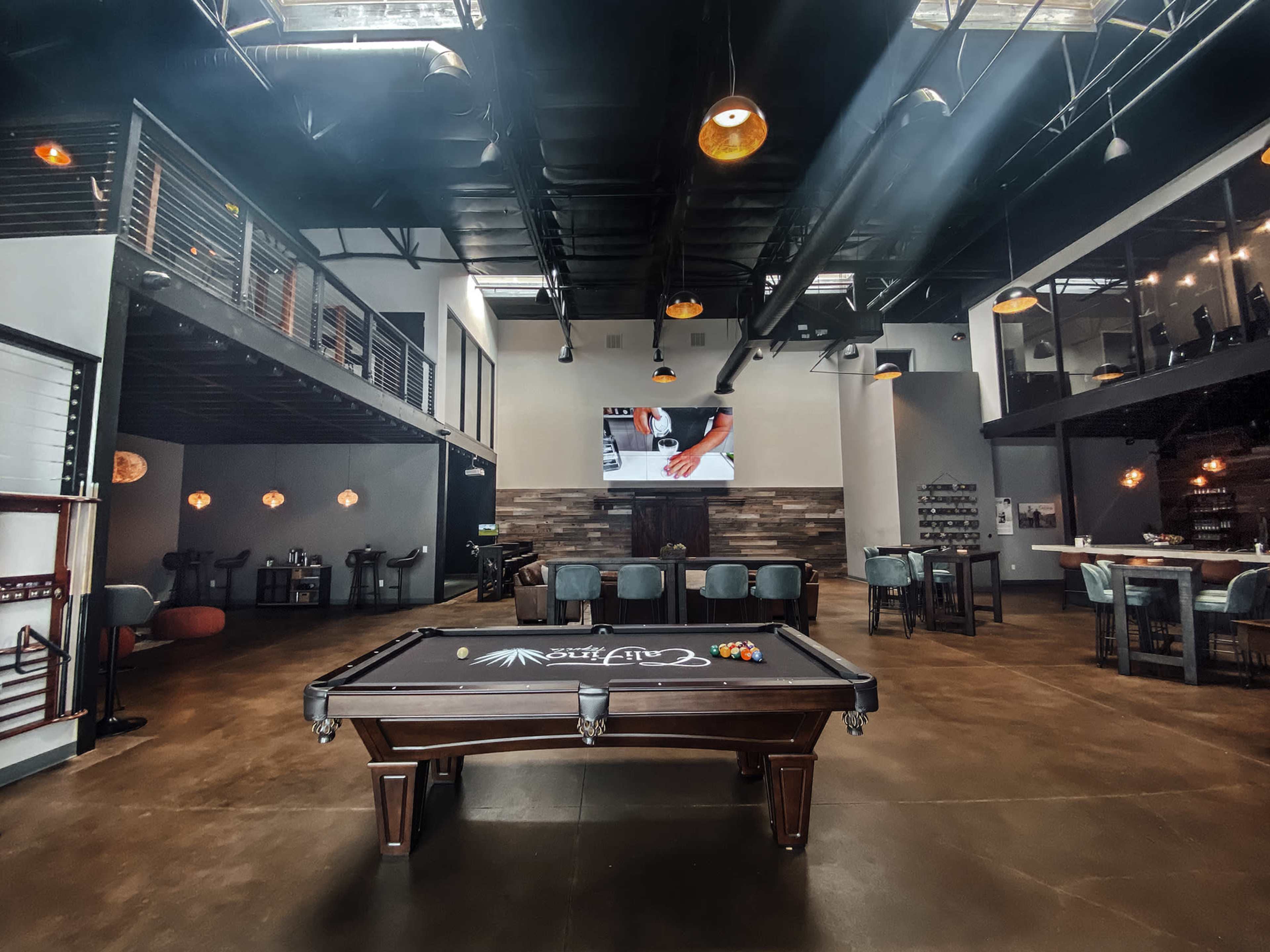 A spacious lounge area featuring a pool table in the foreground, surrounded by modern seating arrangements and a large screen displaying a video in the background.