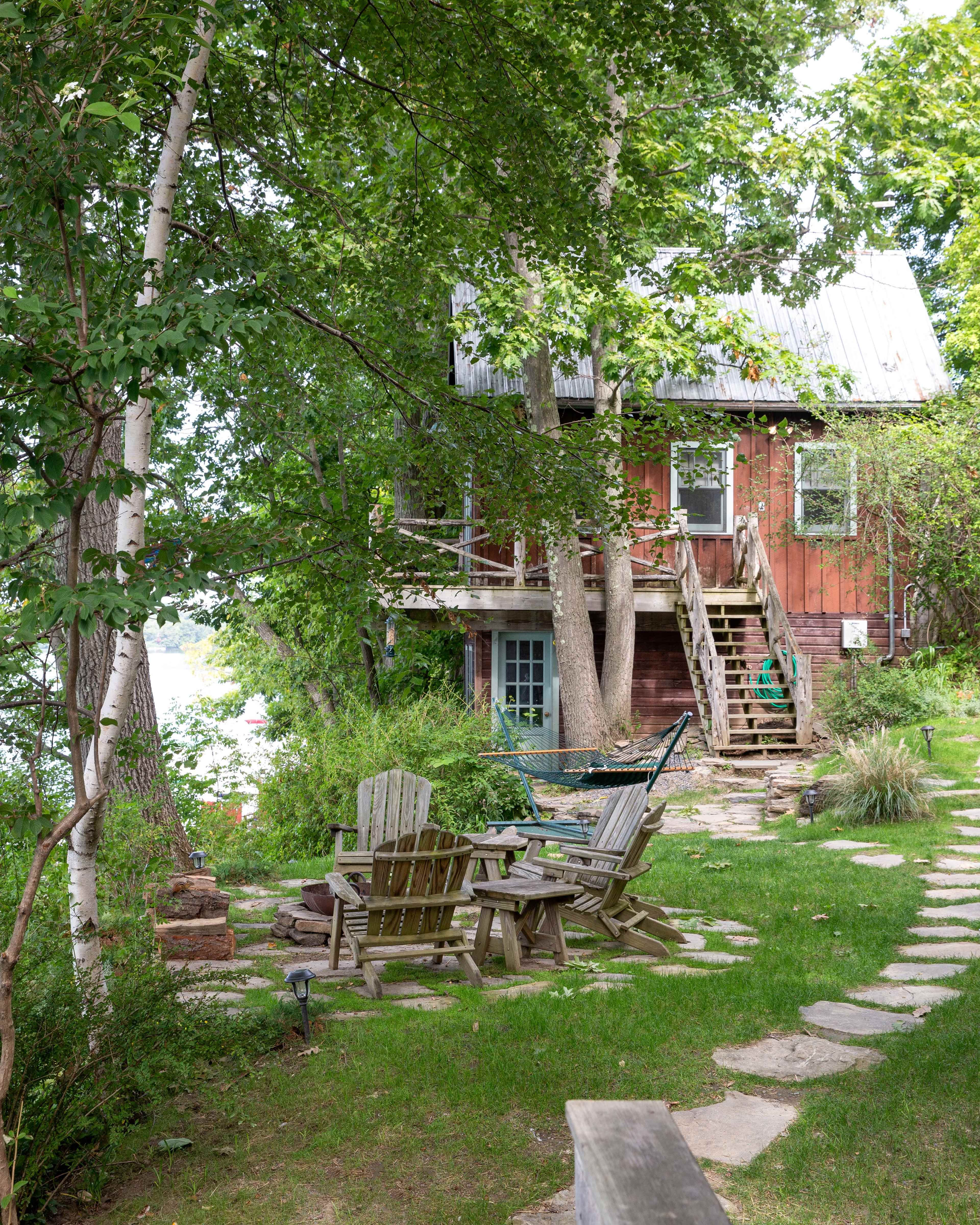 A rustic cabin with a metal roof sits among trees, featuring a stone pathway leading to a seating area with Adirondack chairs.