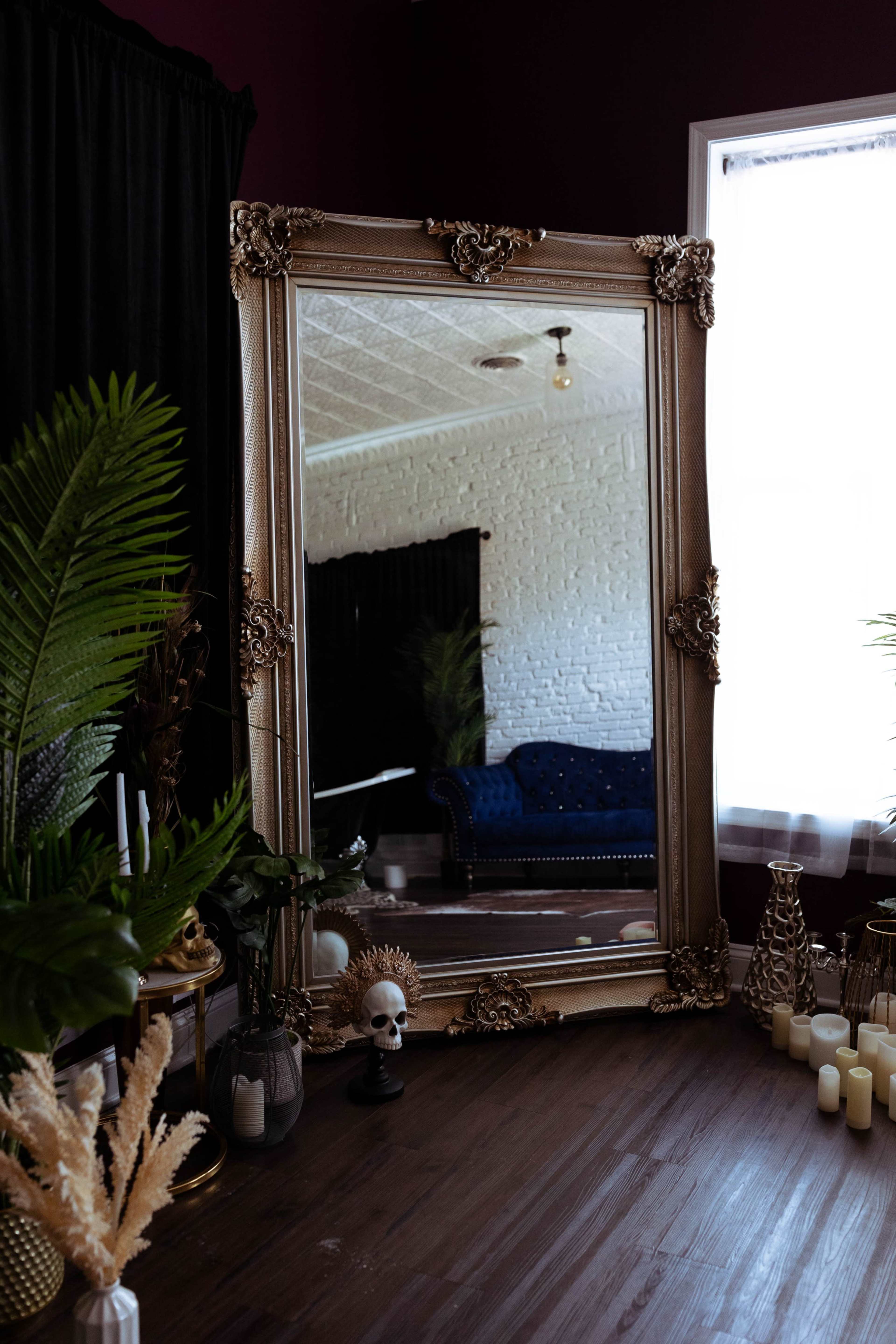 A large ornate mirror reflects a blue couch in a dimly lit room decorated with plants and candles.