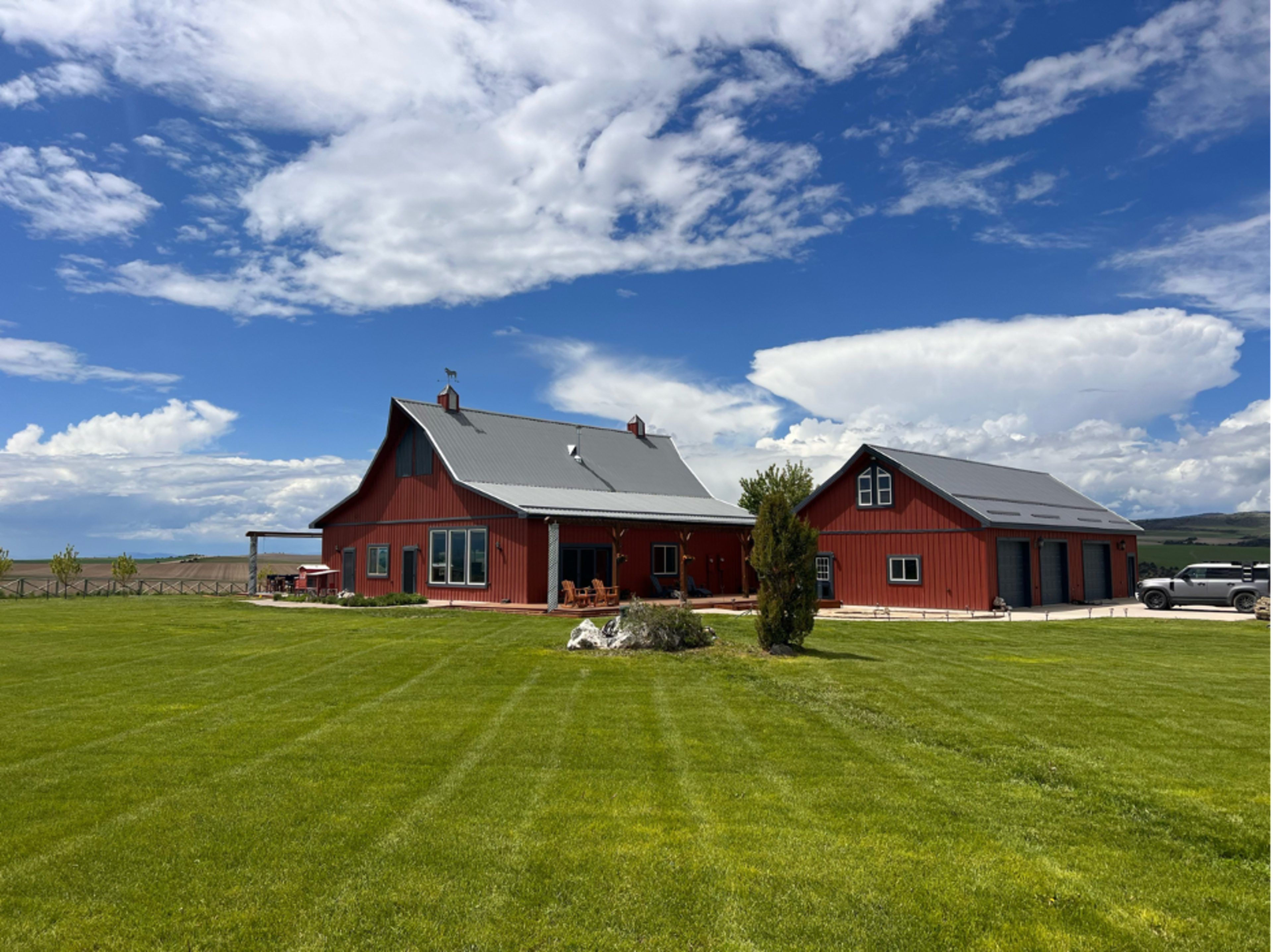 A red barn-style house and adjacent garage sit on a grassy lawn under a partly cloudy blue sky.