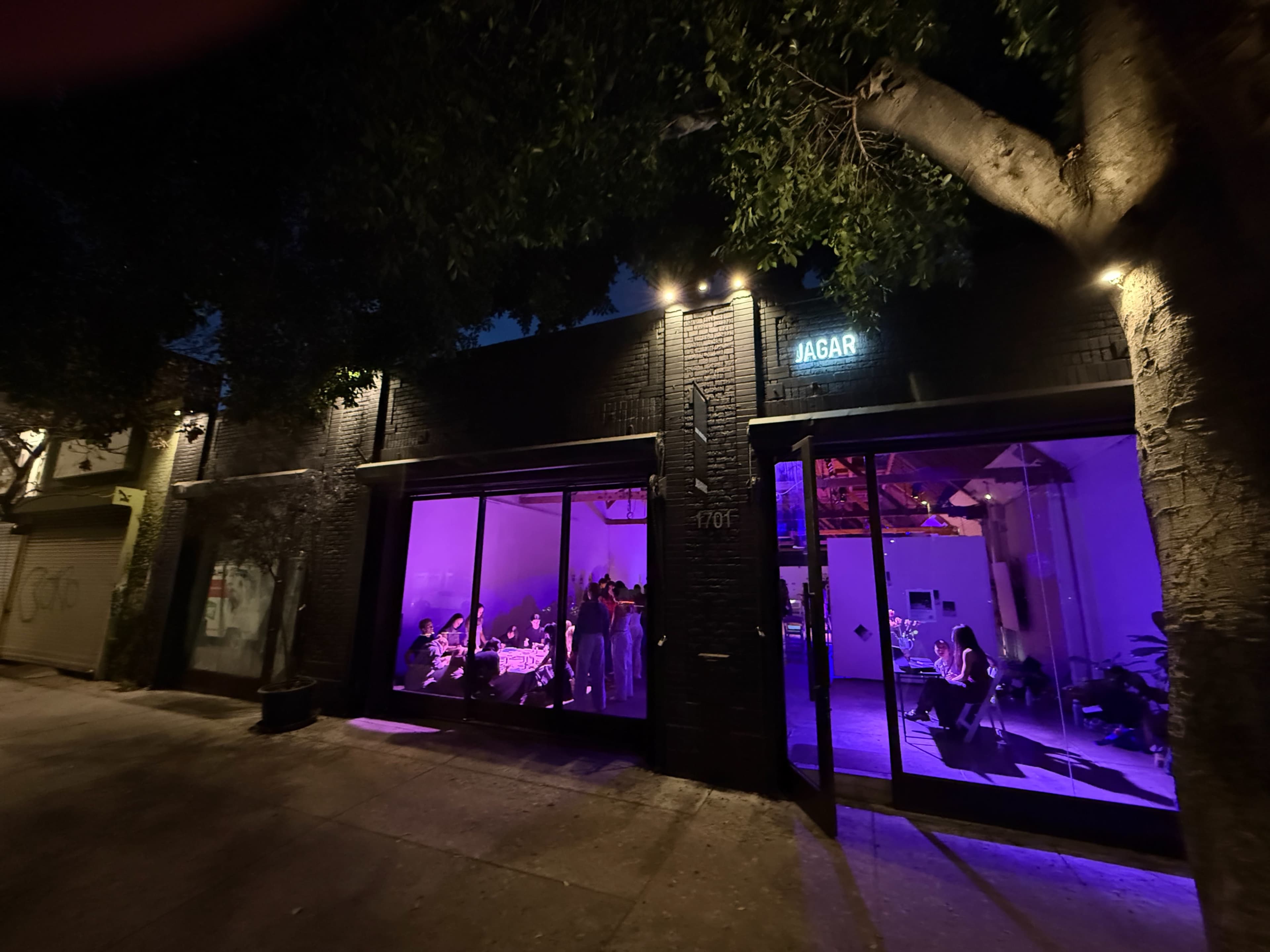 The image shows a dimly lit venue with large glass windows illuminated in purple light, where groups of people are gathered around tables inside.