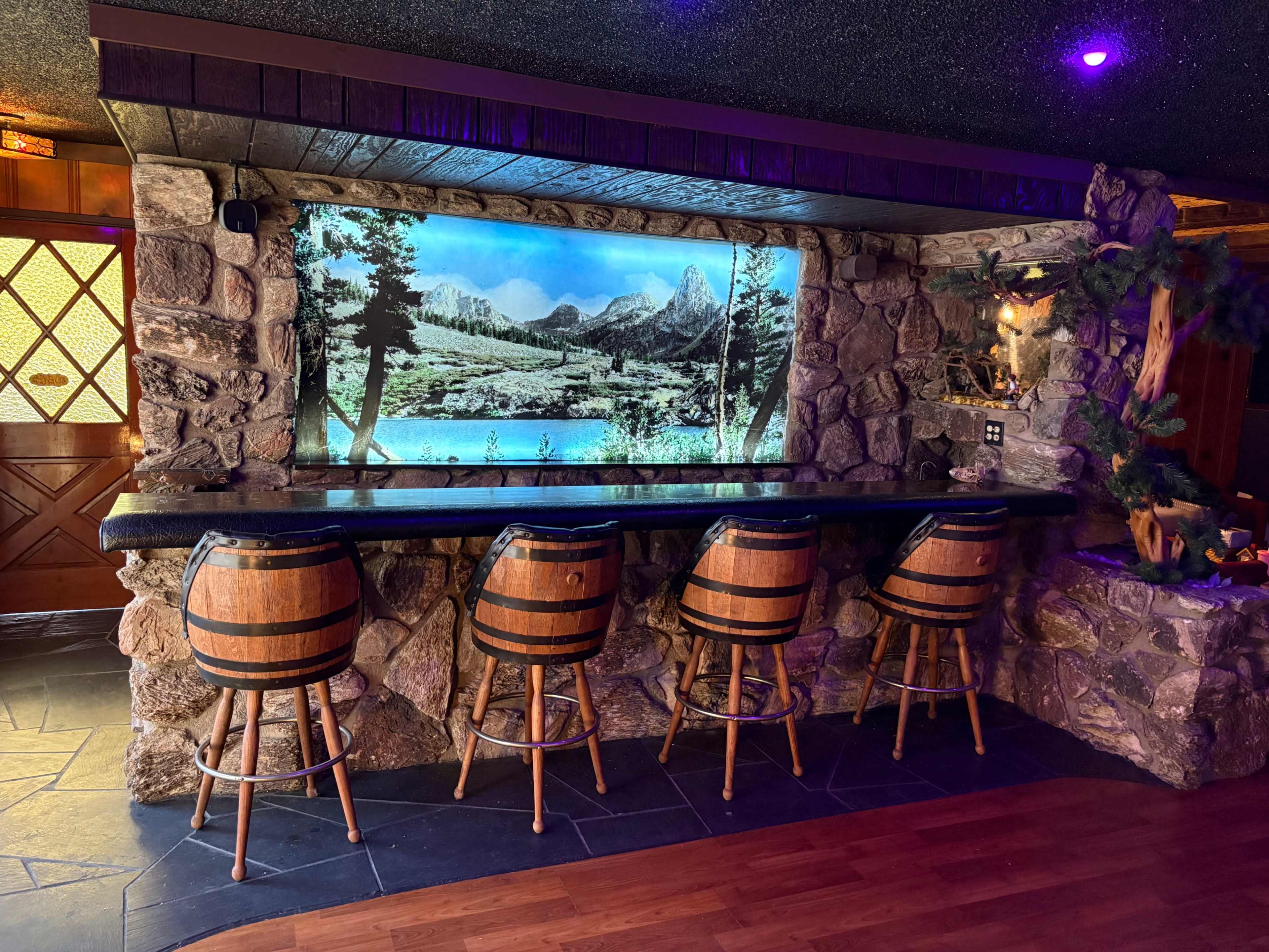 The image shows a rustic bar area with wooden barrel-style stools and a large screen displaying a scenic mountain landscape framed by a stone wall.