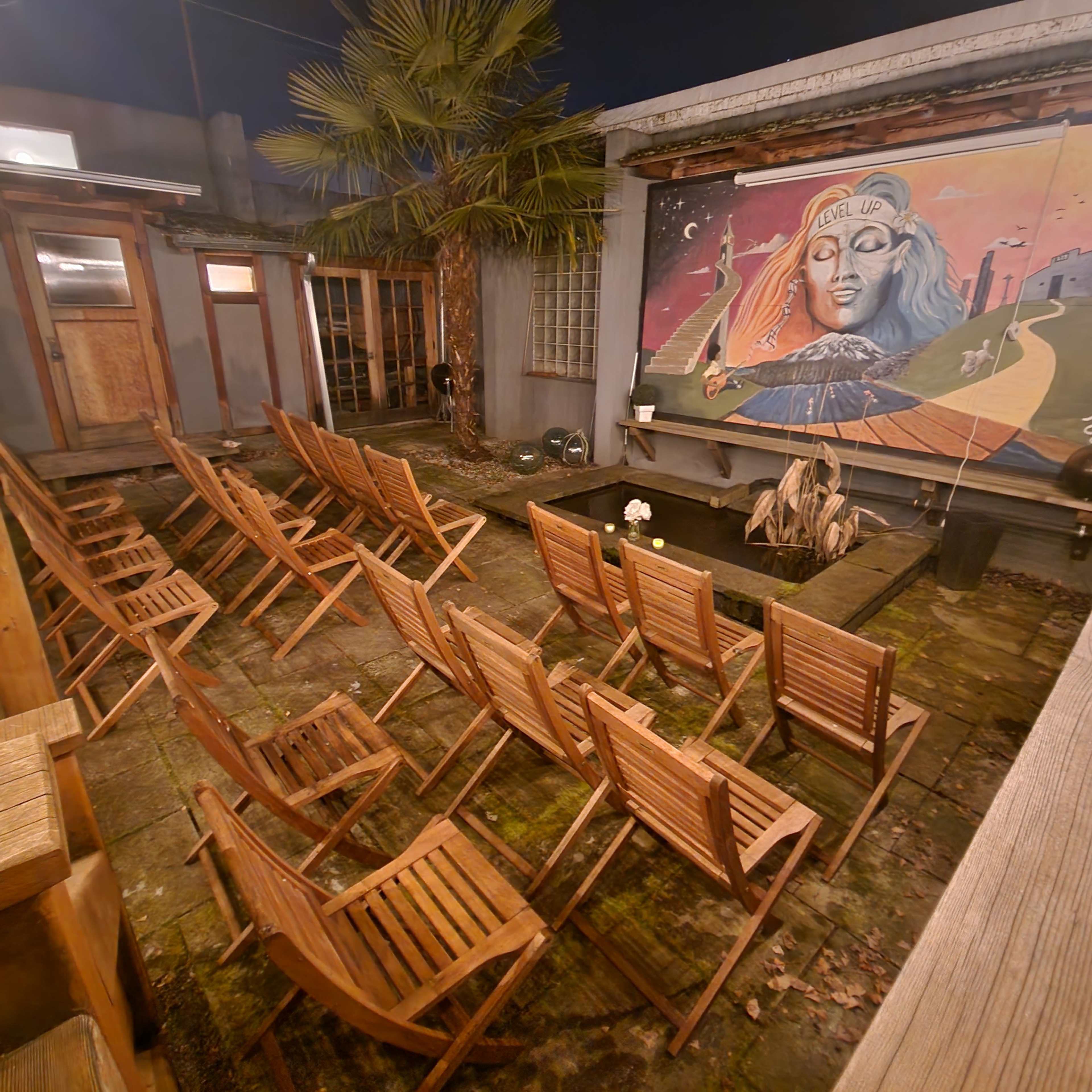 A courtyard with wooden chairs facing a mural of a woman and a small water feature.
