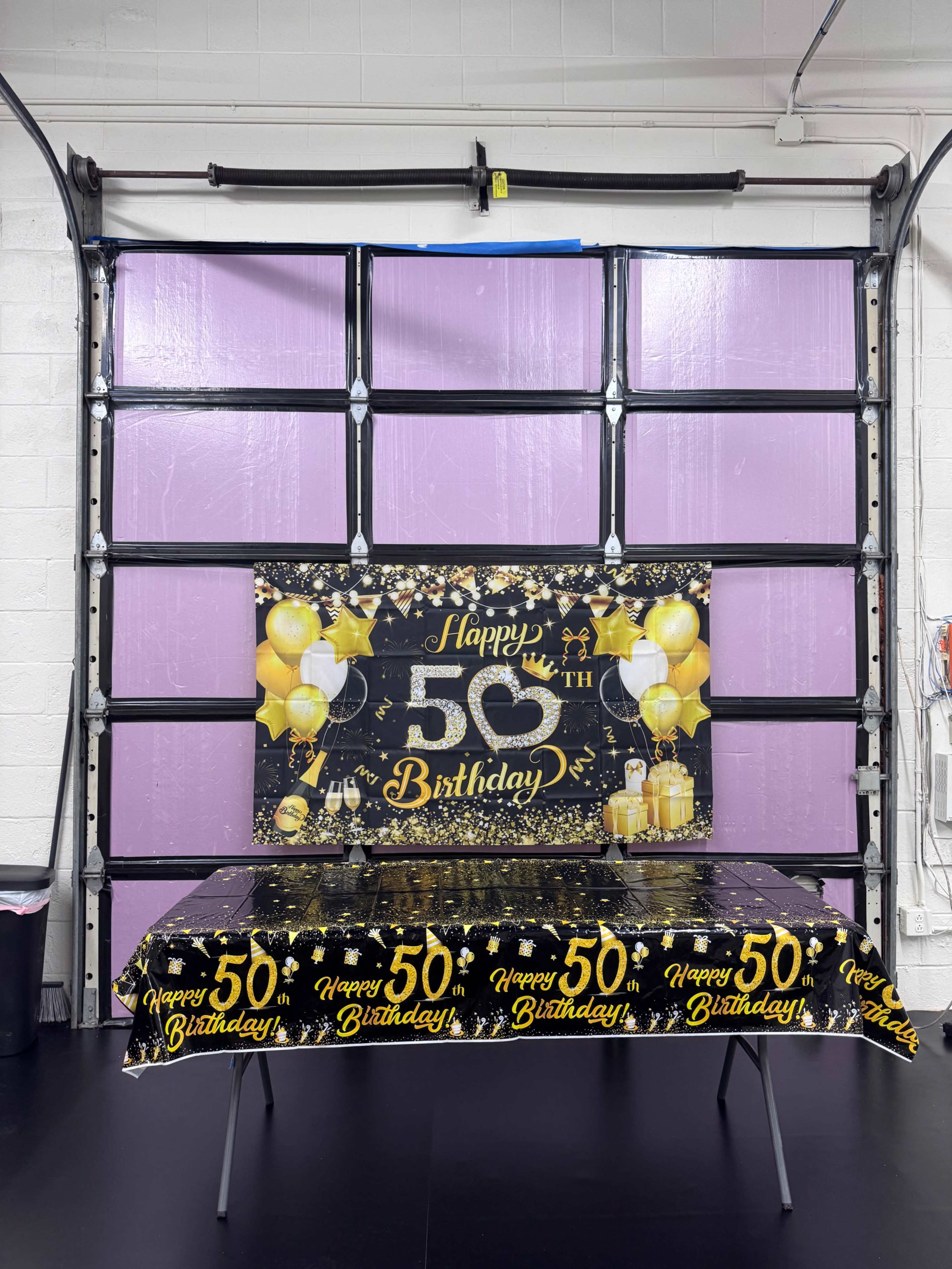 The image shows a decorated table featuring a black and gold birthday banner and tablecloth, set against a garage door with purple panels.