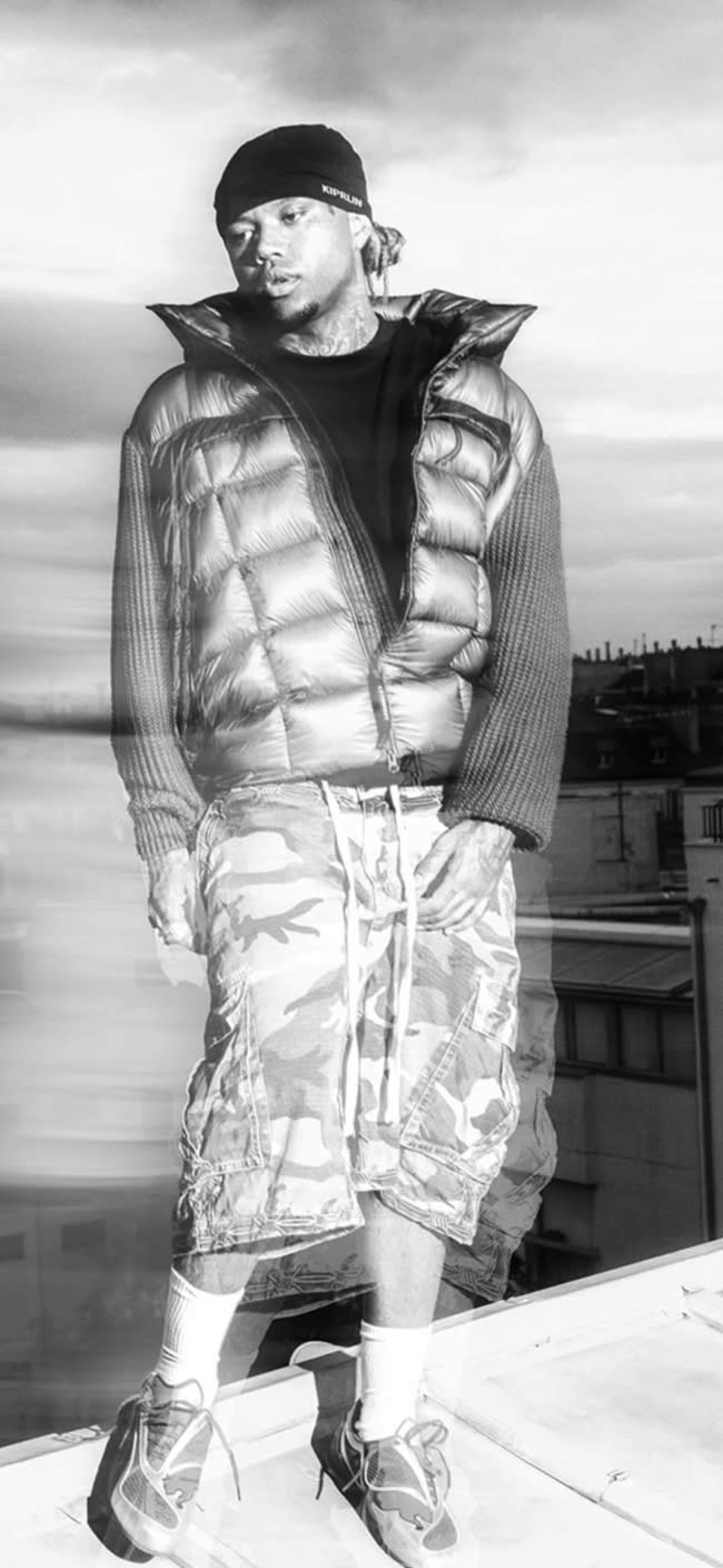 A person wearing a puffy vest and camo shorts stands on a rooftop against a cloudy sky.