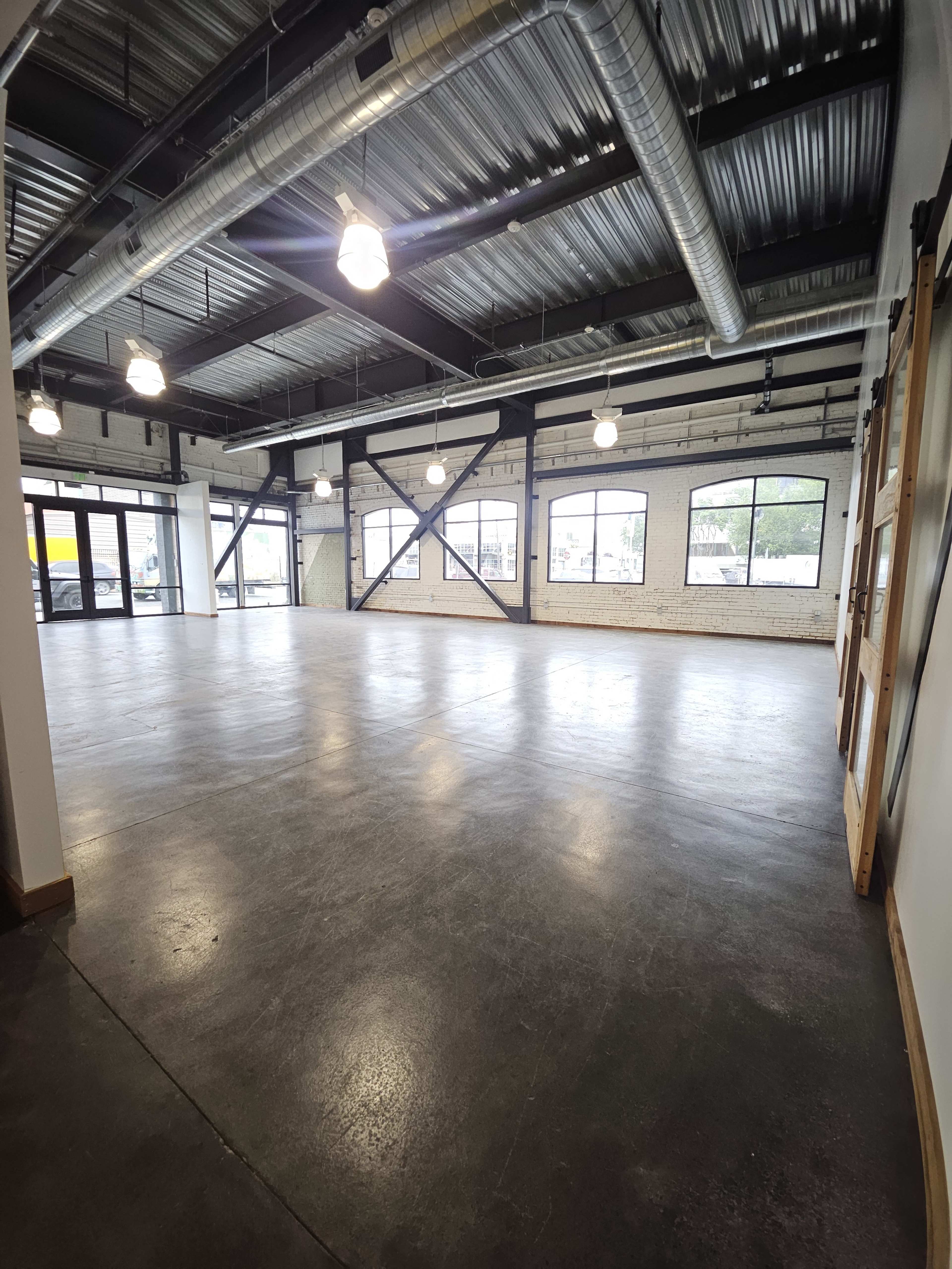 An empty industrial-style interior space with concrete flooring, exposed beams, and large windows.