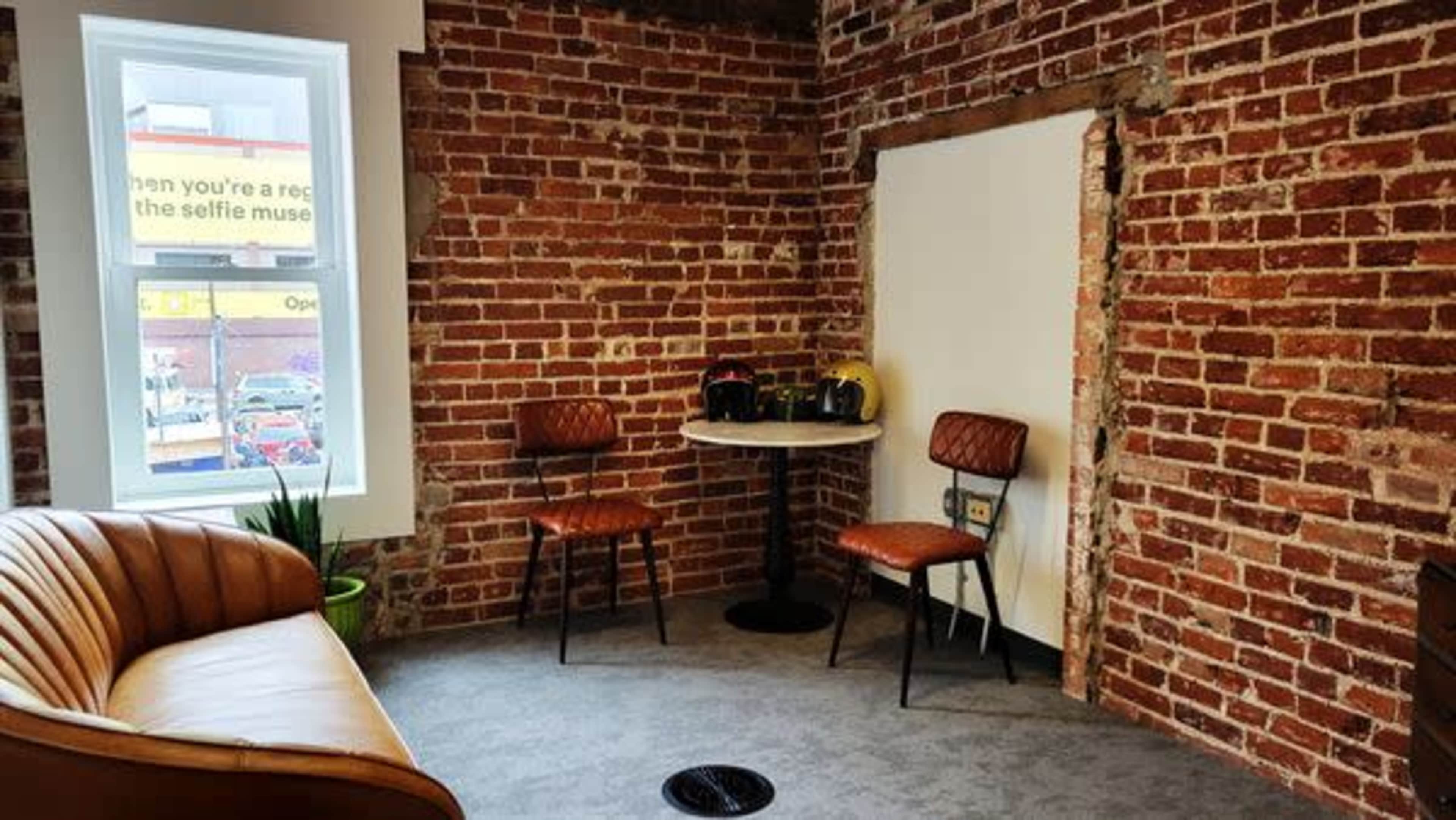 The image shows a small room with exposed brick walls, a round table, two chairs, a brown leather couch, and a window revealing a city street outside.