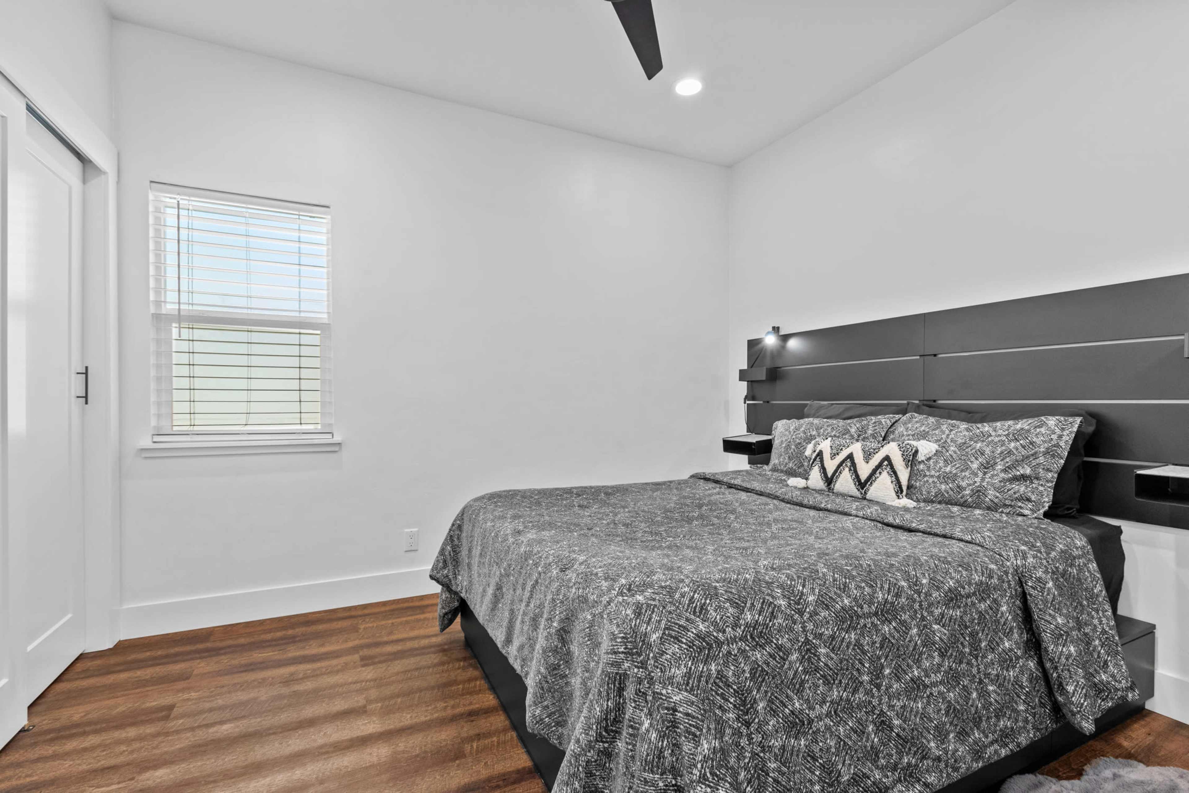 The image shows a modern bedroom with a bed covered in a patterned gray blanket and a minimalist headboard, accompanied by built-in side tables and a window allowing natural light.