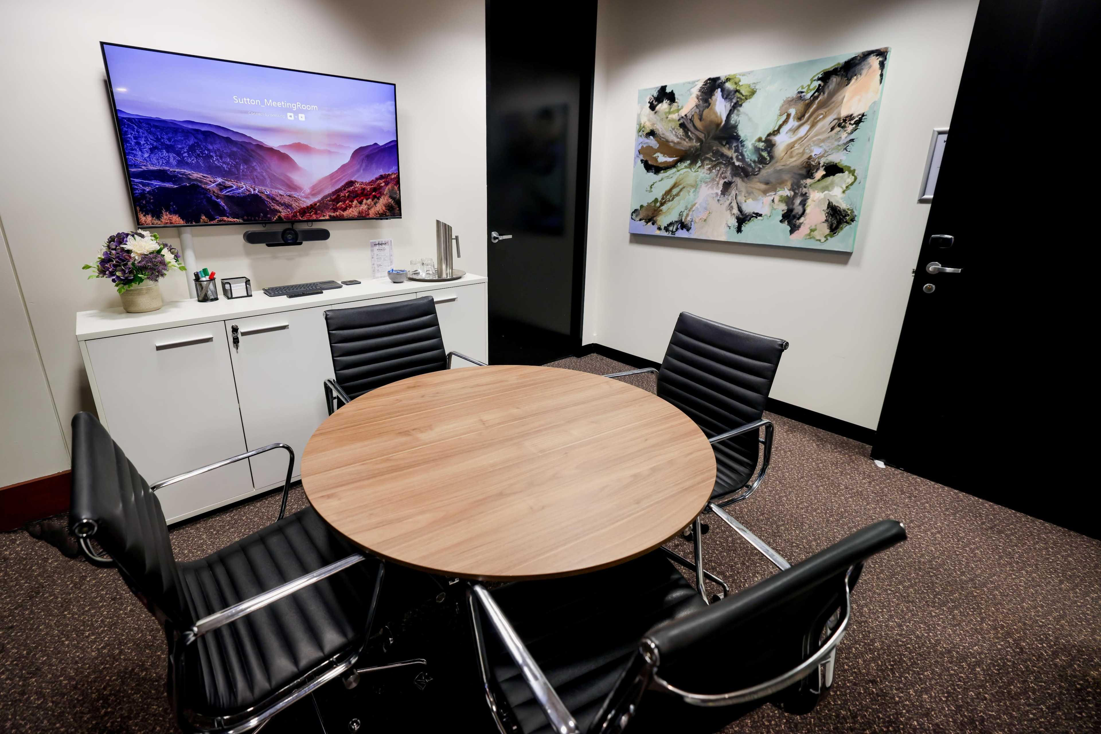 The image shows a small meeting room with a round wooden table, four black leather chairs, a wall-mounted television, and abstract art on the wall.