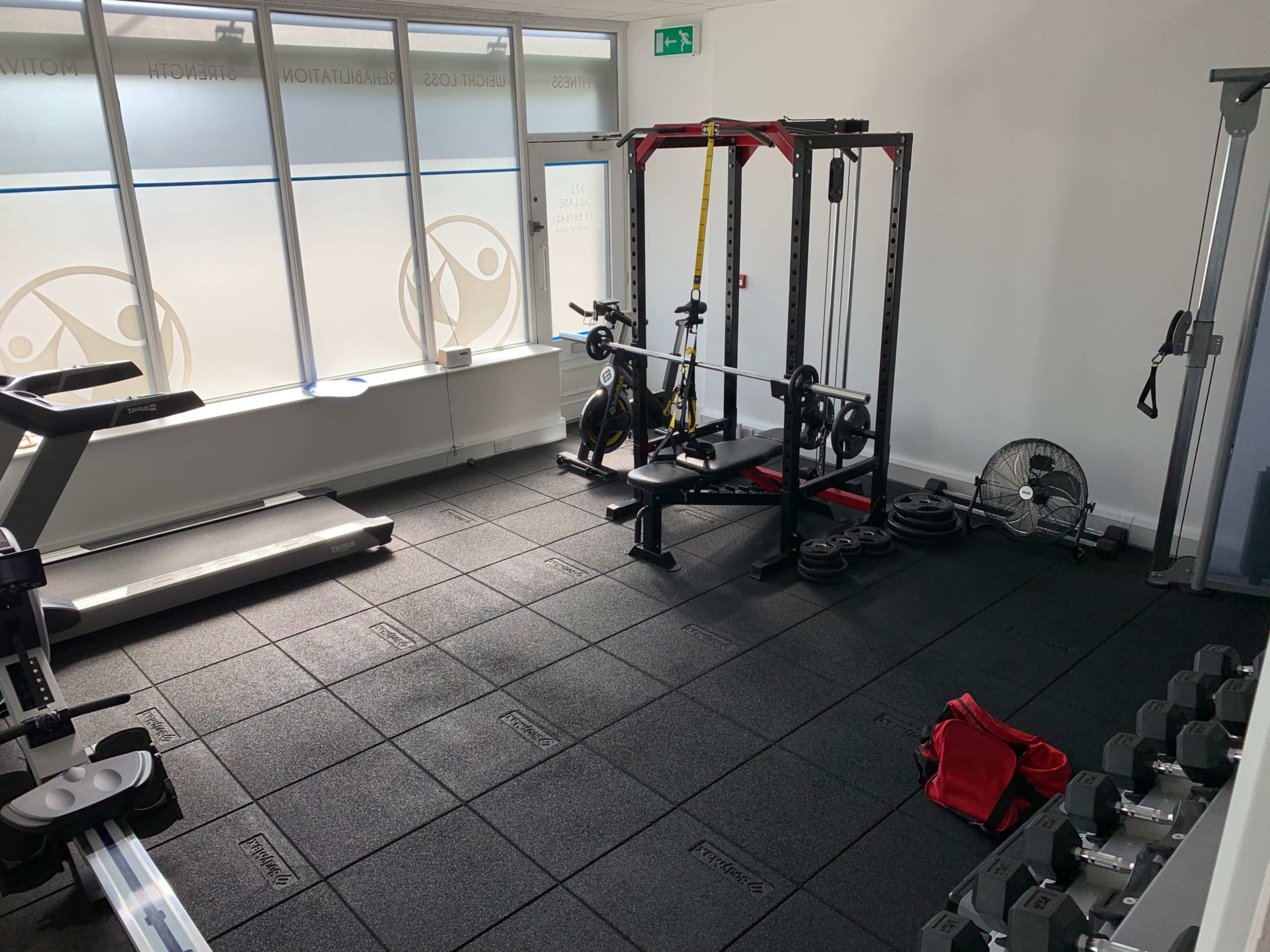 The image shows a fitness area equipped with various gym equipment, including a barbell rack, dumbbells, a treadmill, and a fan, on a black rubber flooring surface.