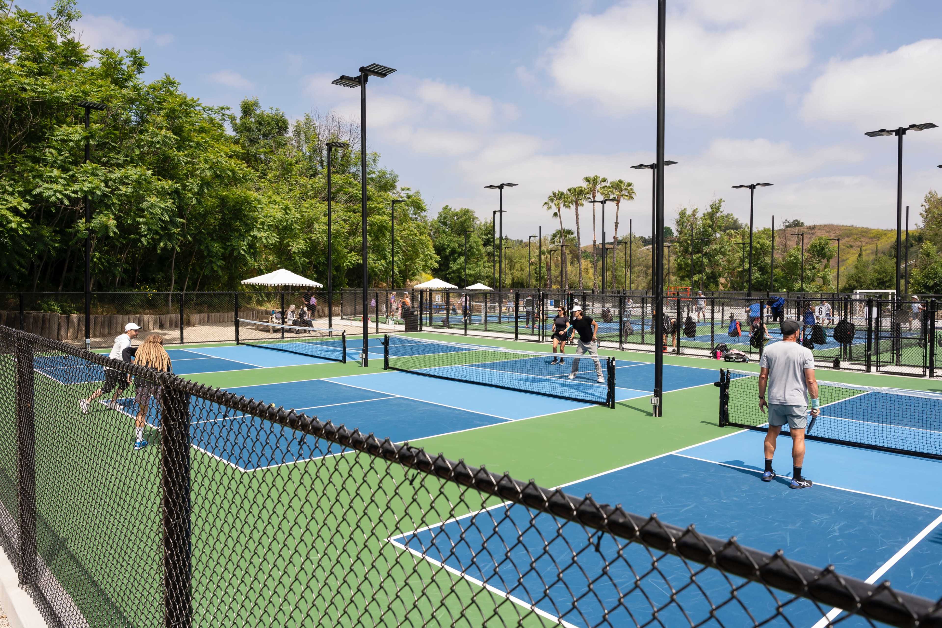 A vibrant outdoor pickleball complex with multiple courts, players engaged in games, and spectators sitting under umbrellas in a landscaped area.