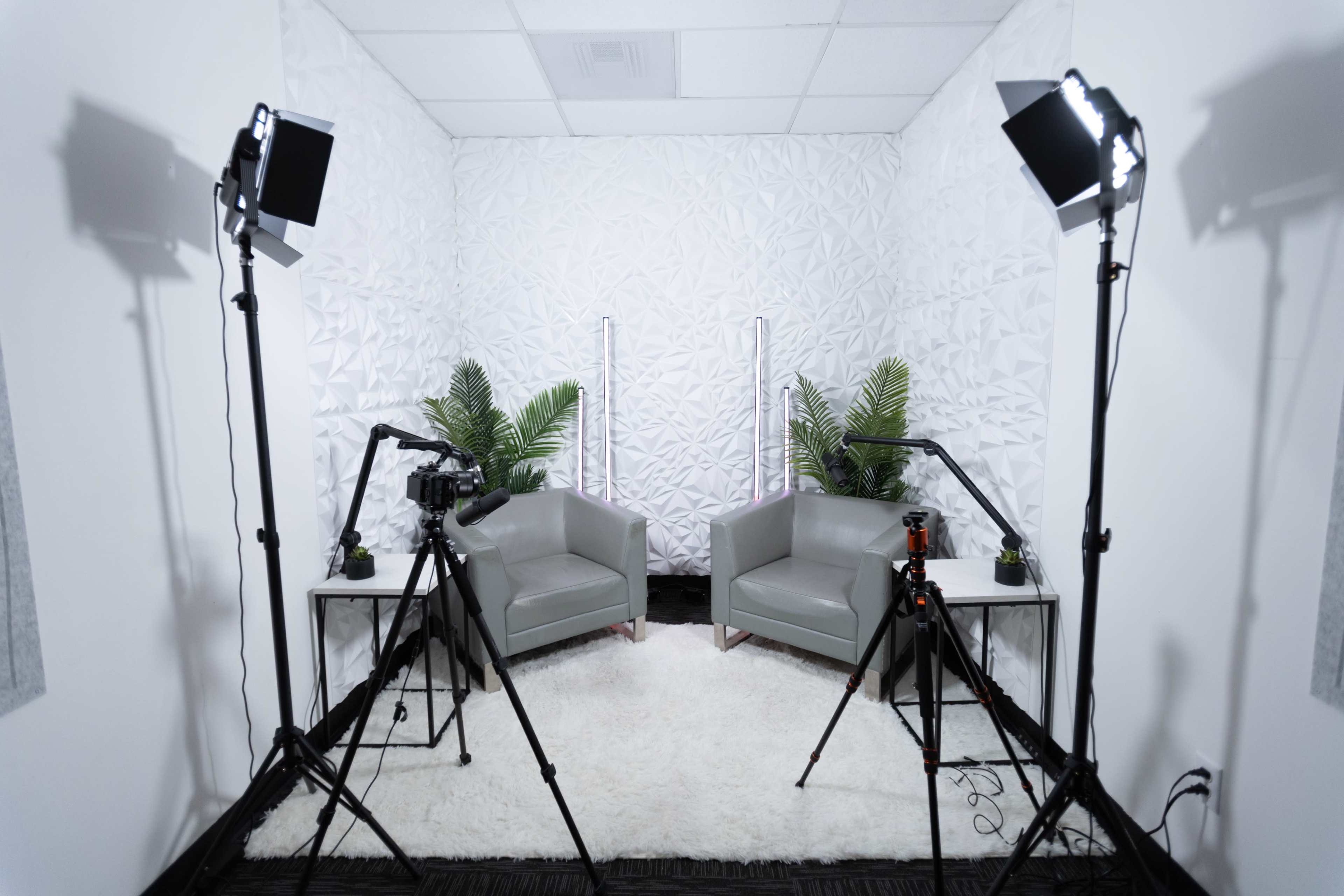 A studio setup with two grey chairs, a white textured backdrop, and lighting equipment positioned around a white rug.