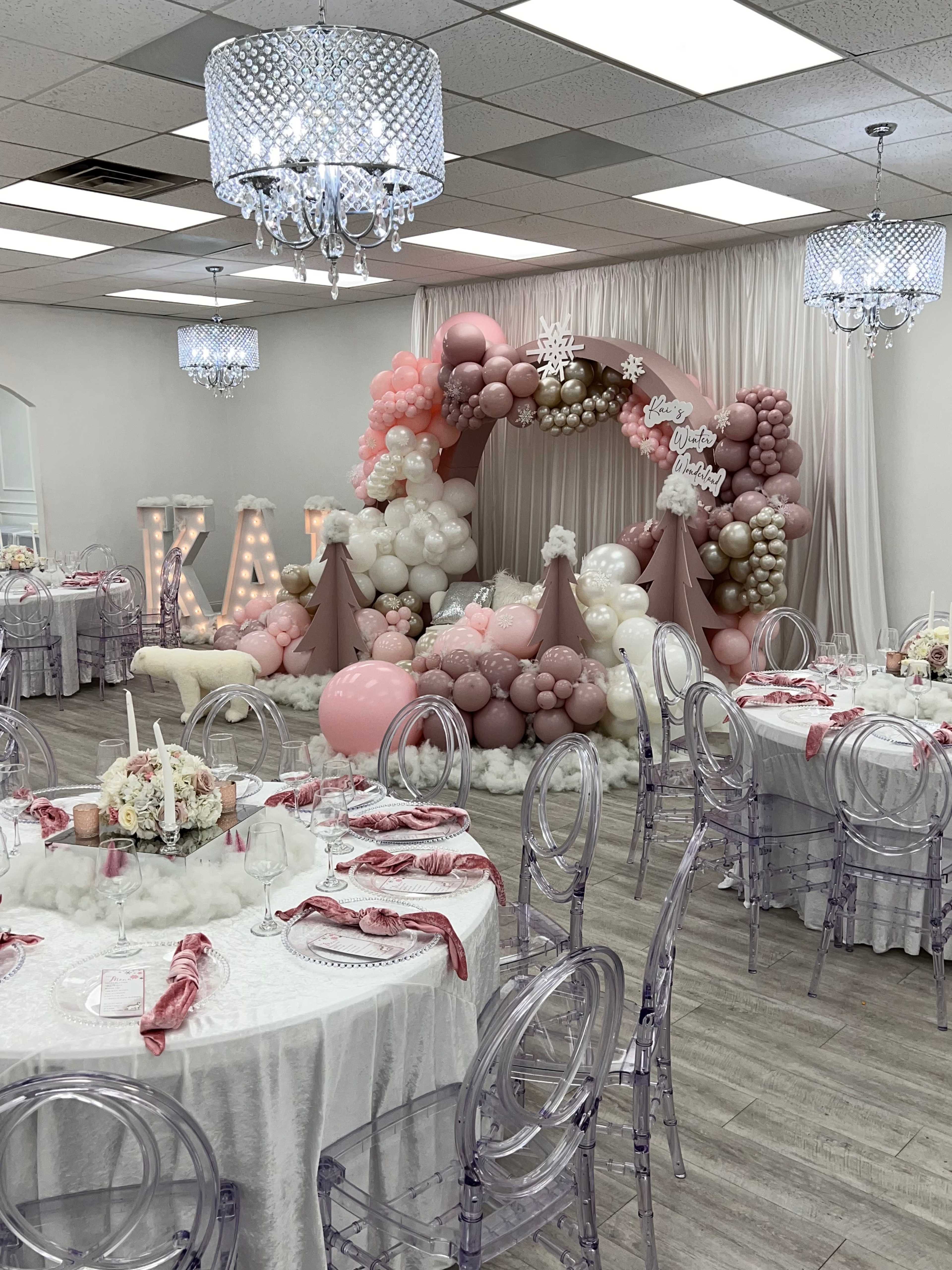 A decorated event space with elegant tables set for a celebration, featuring a backdrop of balloons in shades of pink and white.