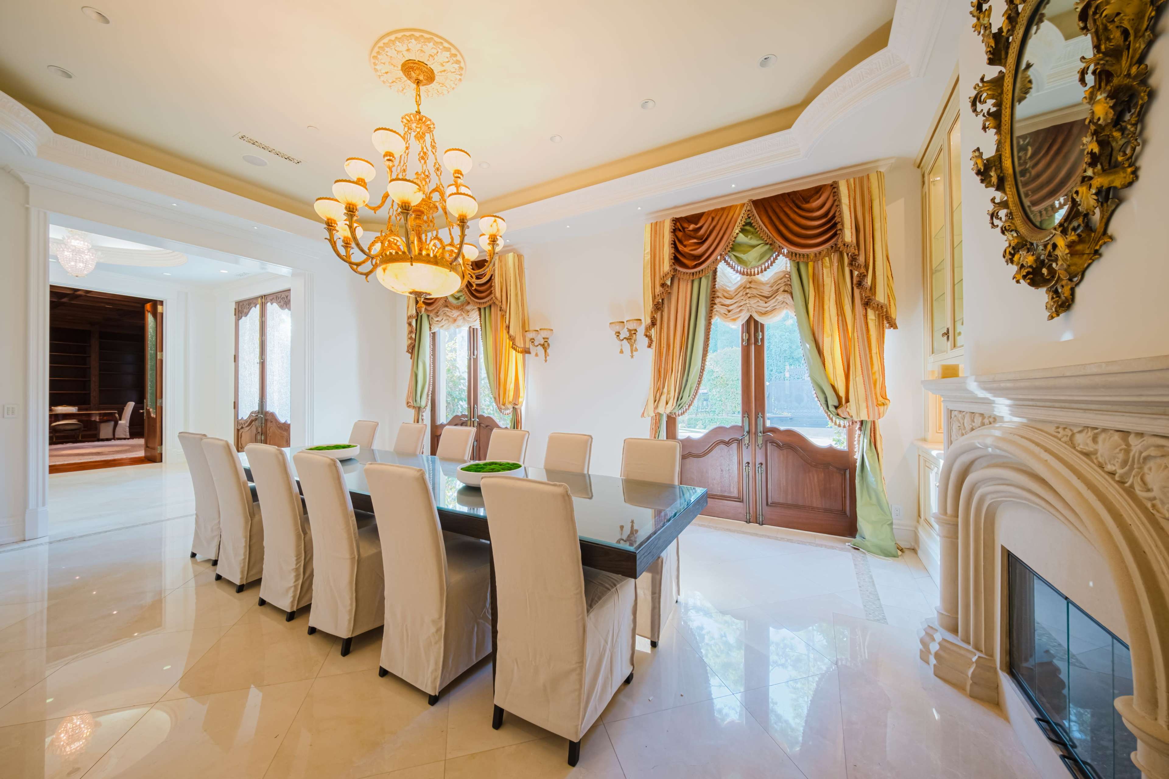 A spacious dining room features a long table surrounded by ten upholstered chairs, with a chandelier overhead and large windows draped with elegant curtains.