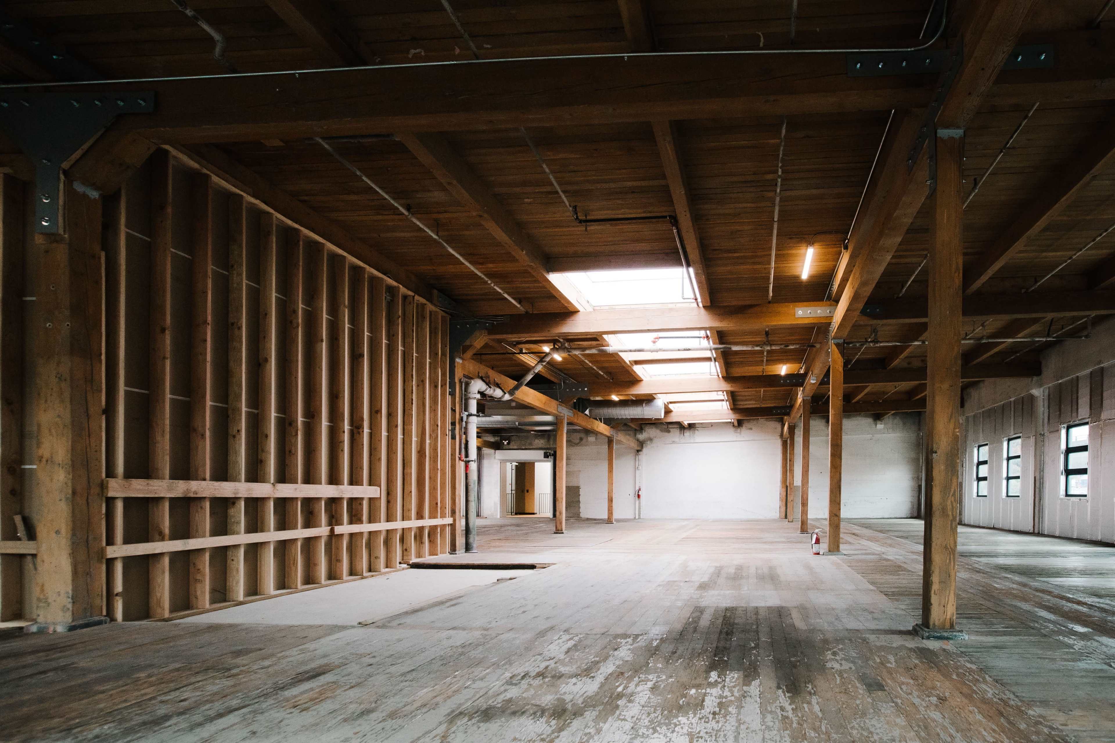 The image shows an empty industrial interior with wooden beams, large windows, and unfinished wooden flooring.