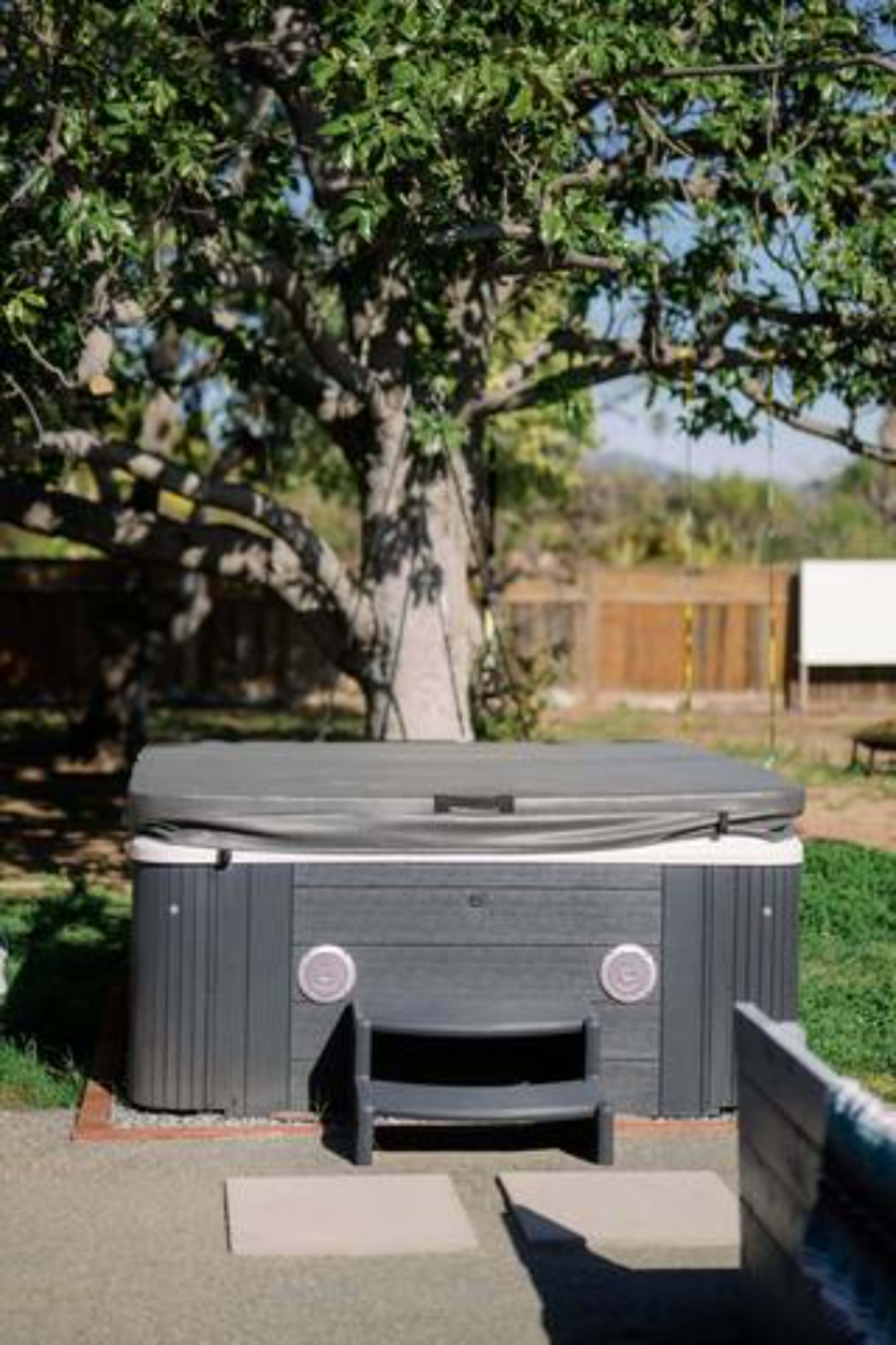 A hot tub is situated on a paved surface beneath a tree in a backyard.