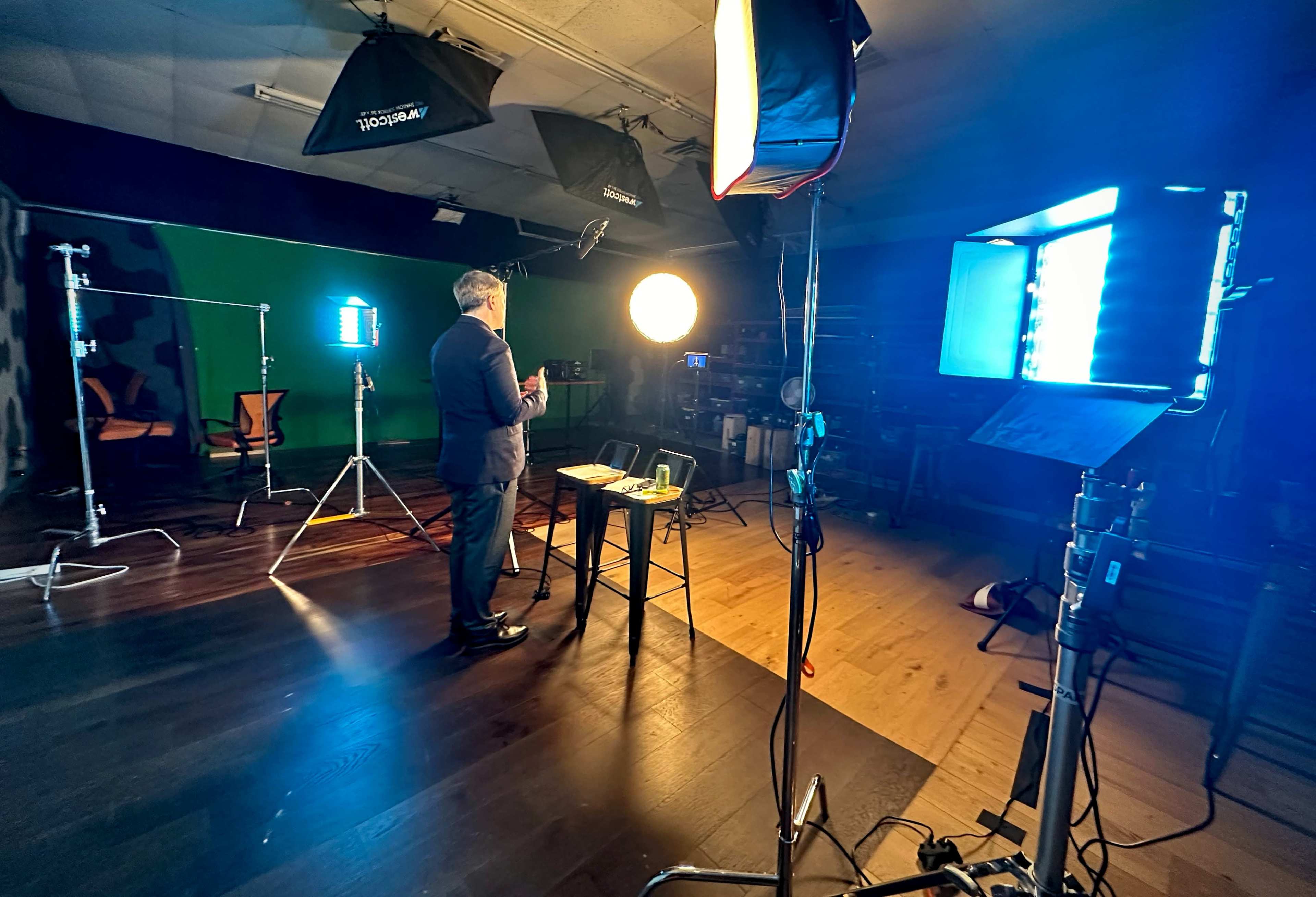 A man stands behind a table in a studio, illuminated by multiple lights and set against a green screen backdrop.