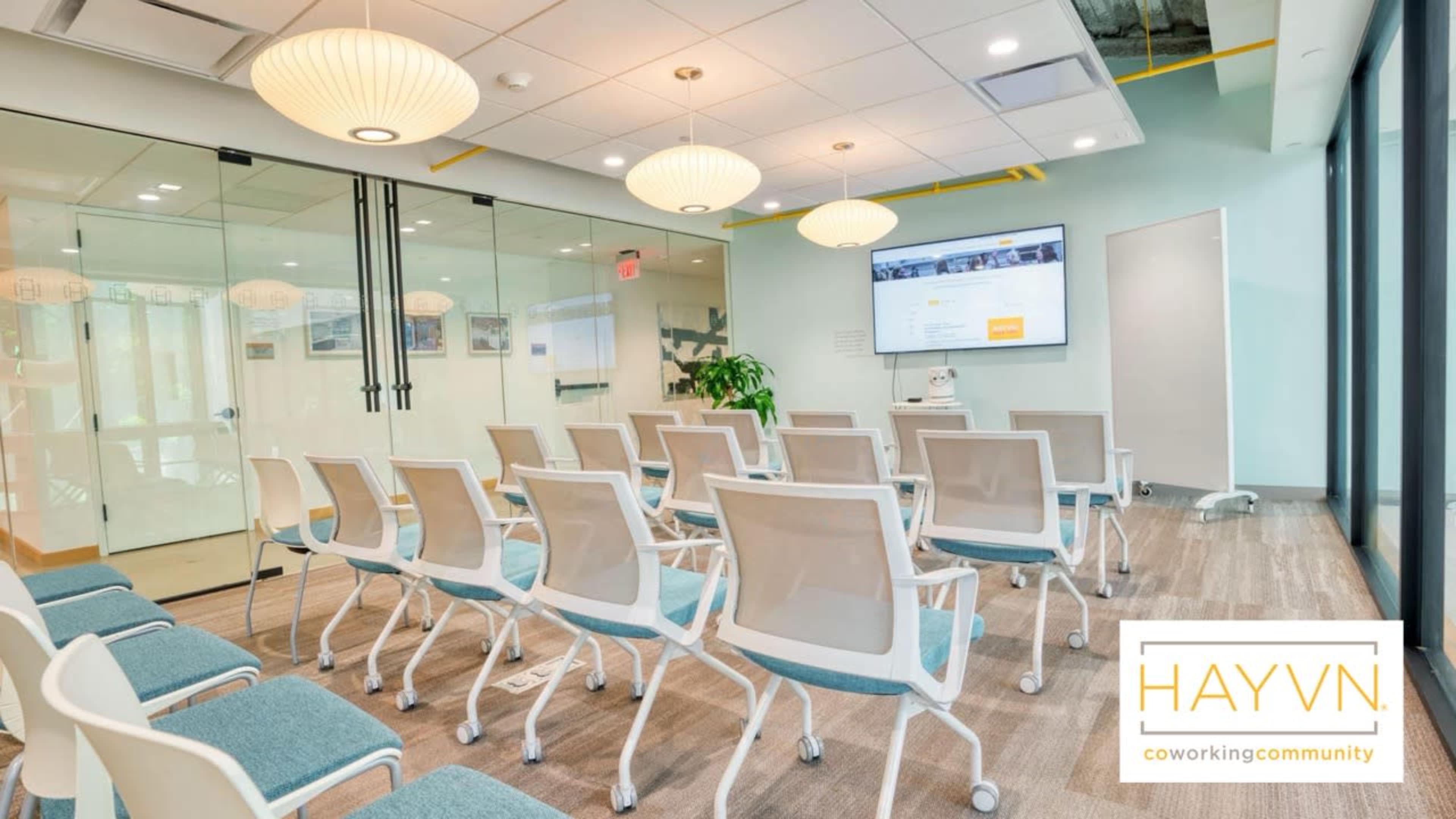 The image shows a modern conference room with rows of white chairs, a large screen projector, and glass walls.