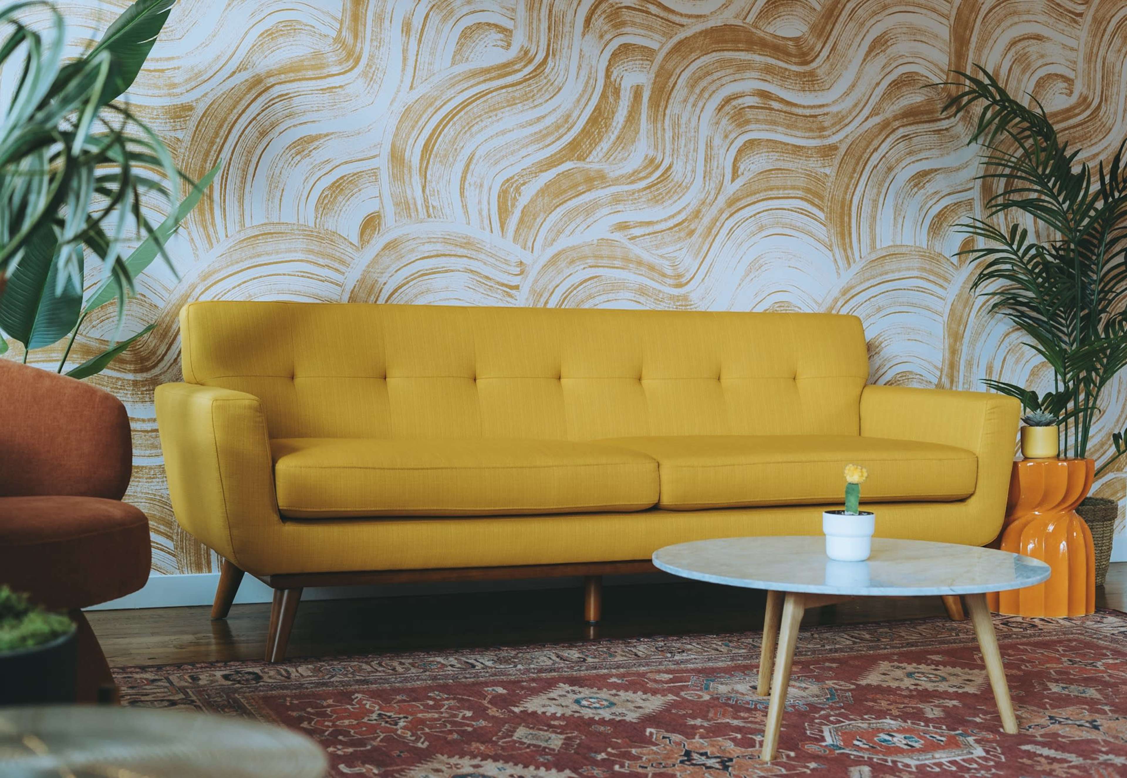 A yellow sofa is positioned in front of a textured, wavy golden wall, accompanied by a round coffee table and potted plants.