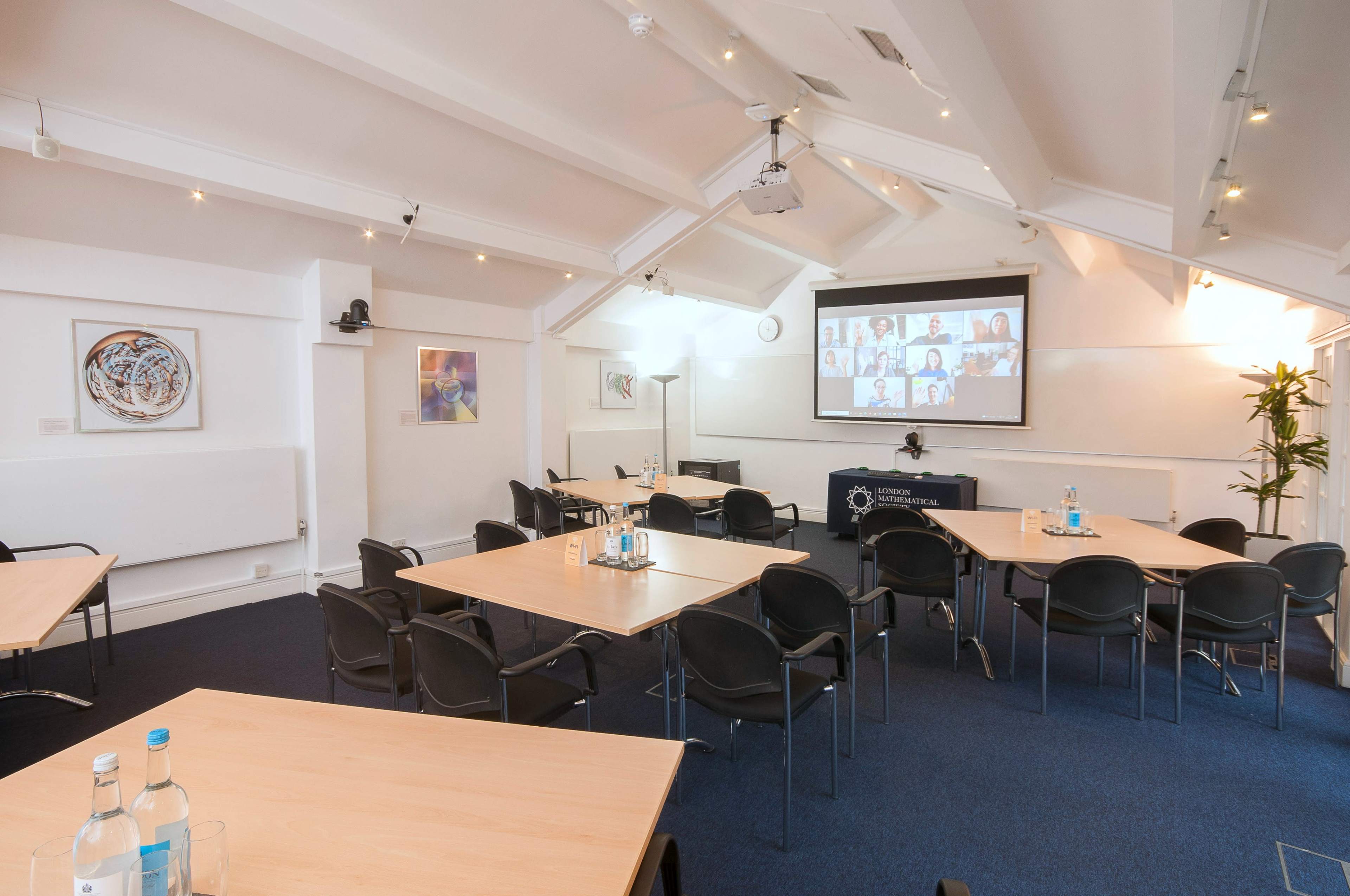 A spacious conference room is set up with multiple tables and chairs, featuring a large screen displaying a video call with participants.