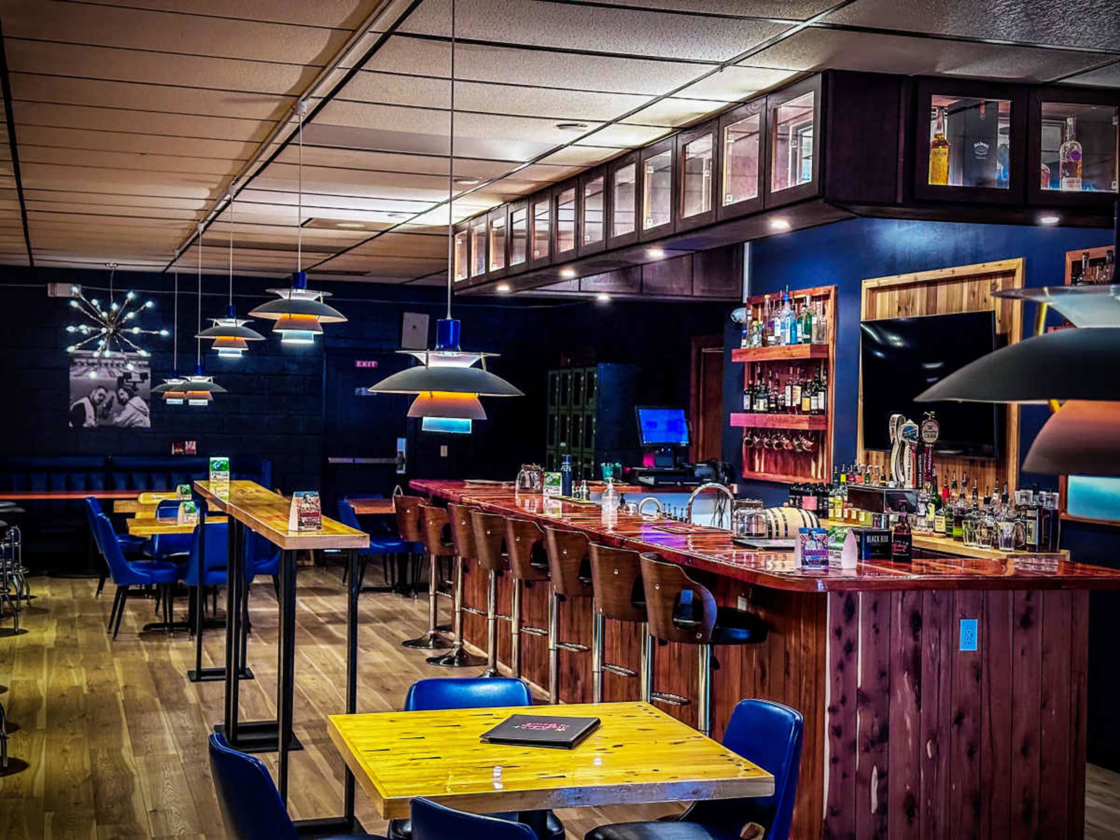 The image shows a modern bar interior with wooden tables, high stools, and a well-stocked bar counter illuminated by pendant lights.