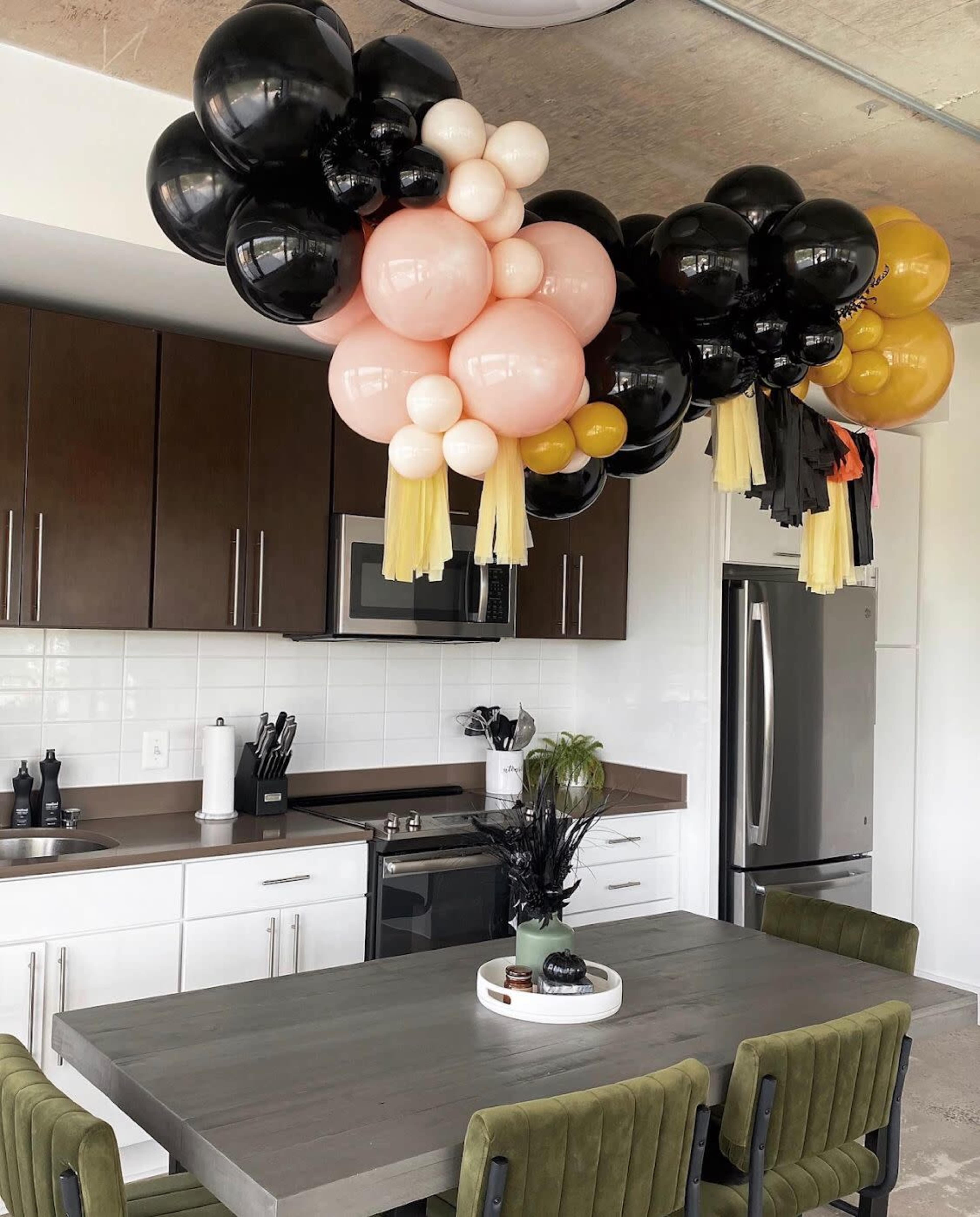 A cluster of balloons in black, pink, white, and gold hangs above a kitchen table with green chairs in a modern kitchen.