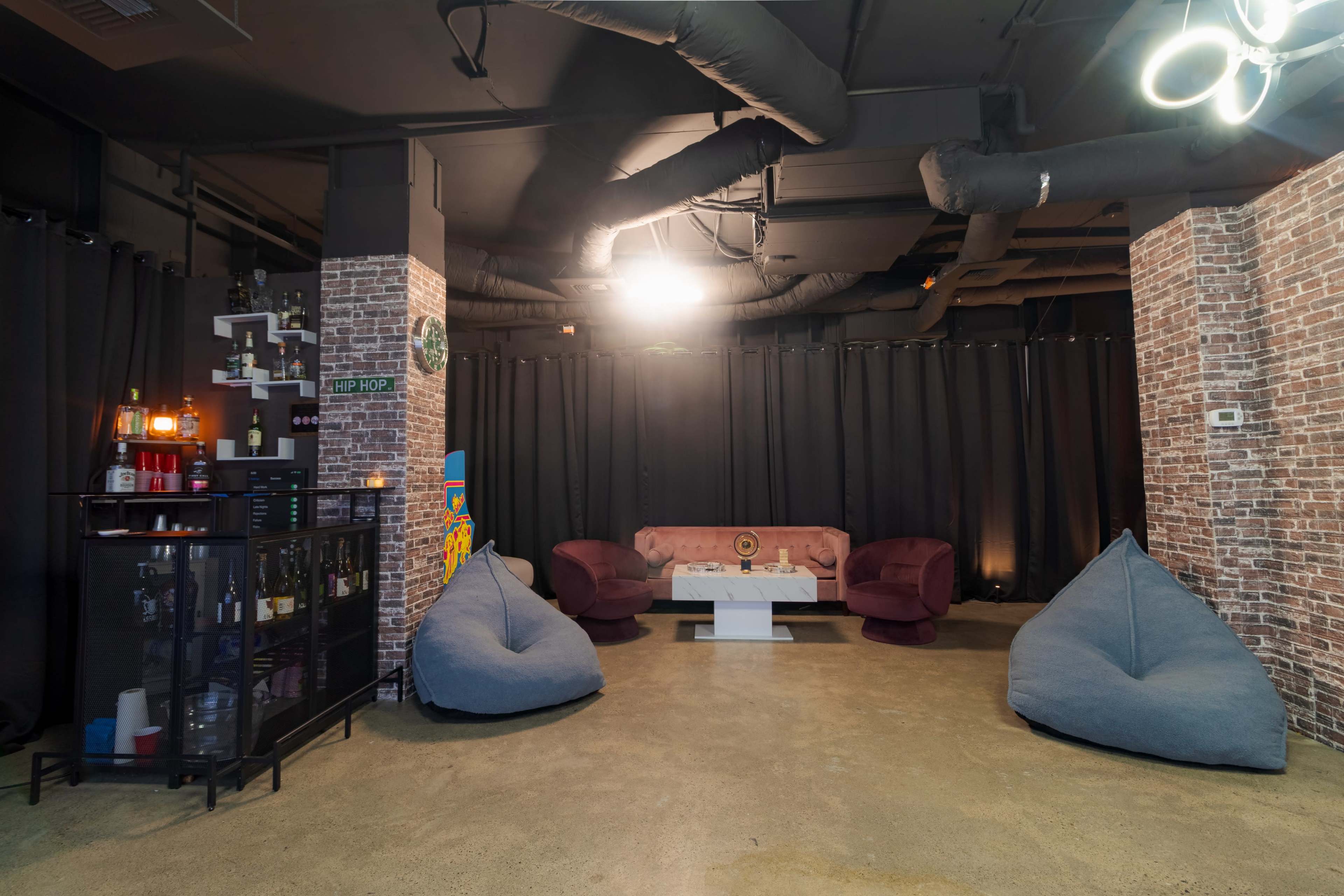 The image shows a lounge area featuring bean bag chairs, a central table, and a sofa, with exposed brick walls and black curtains in the background.