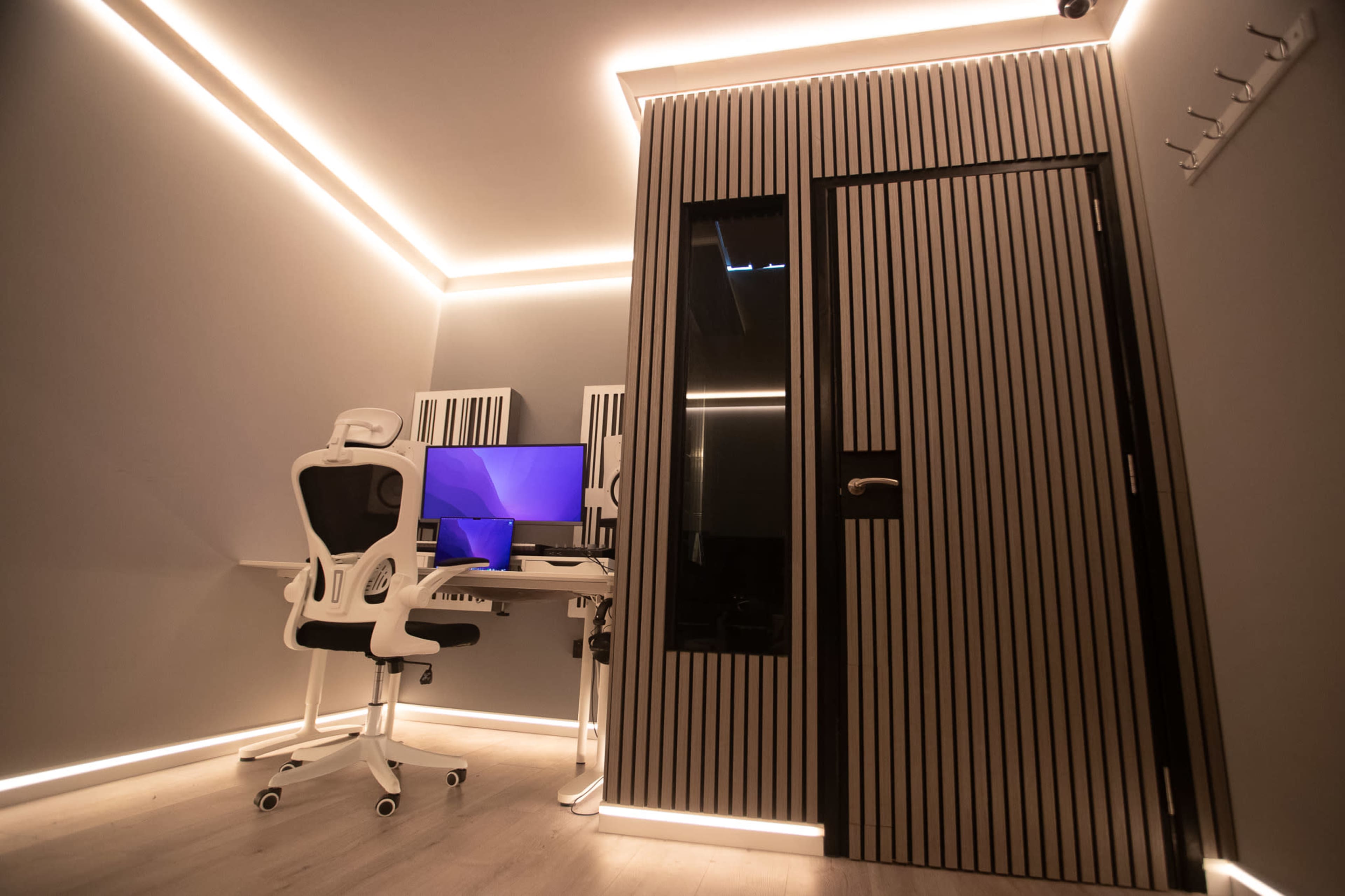 The image shows a modern workspace with a desk set up featuring a computer and an ergonomic chair, alongside a glass-doored room with striped walls illuminated by soft lighting.
