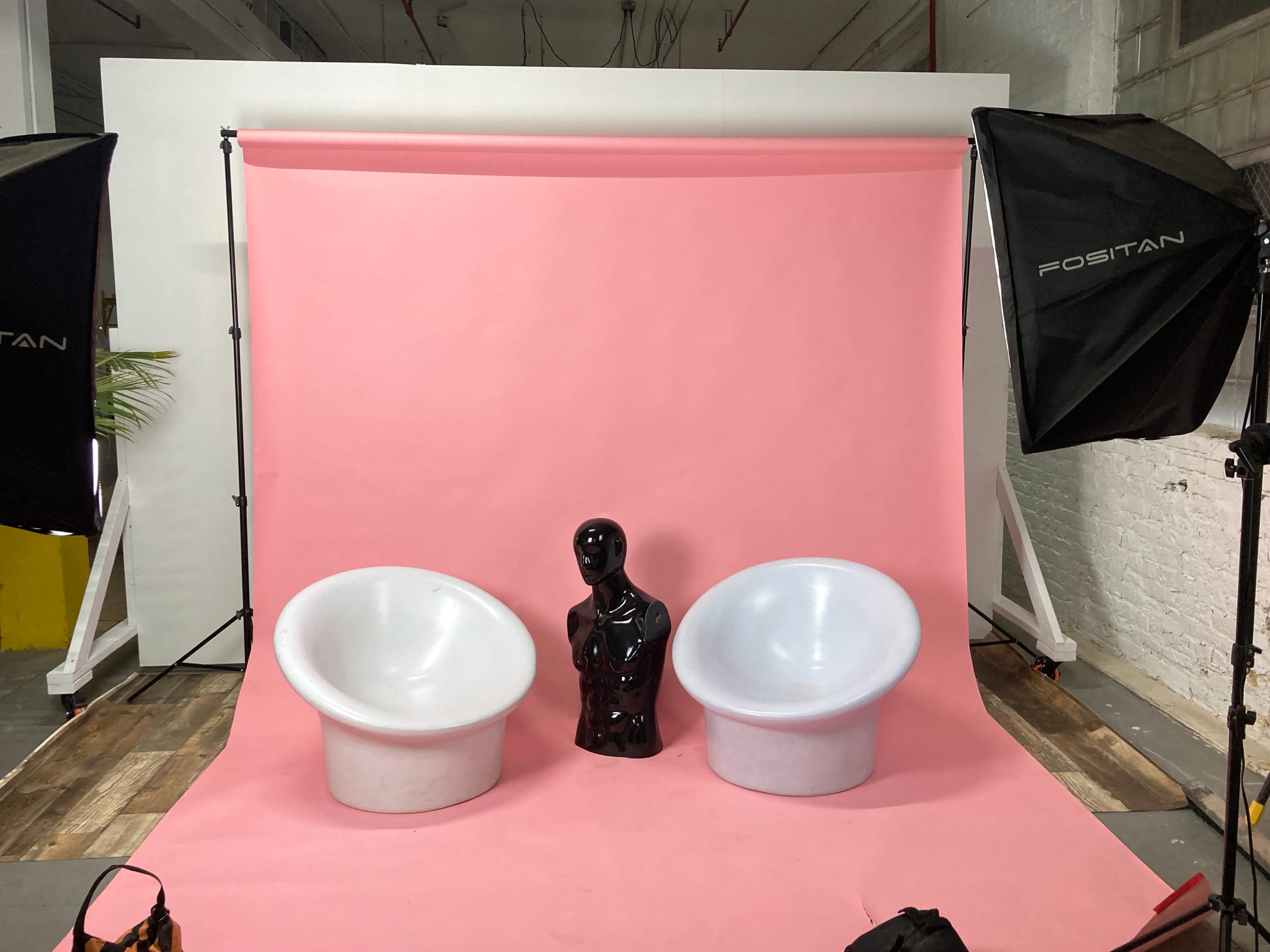 Two white round chairs flanking a black mannequin against a pink backdrop in a photo studio.
