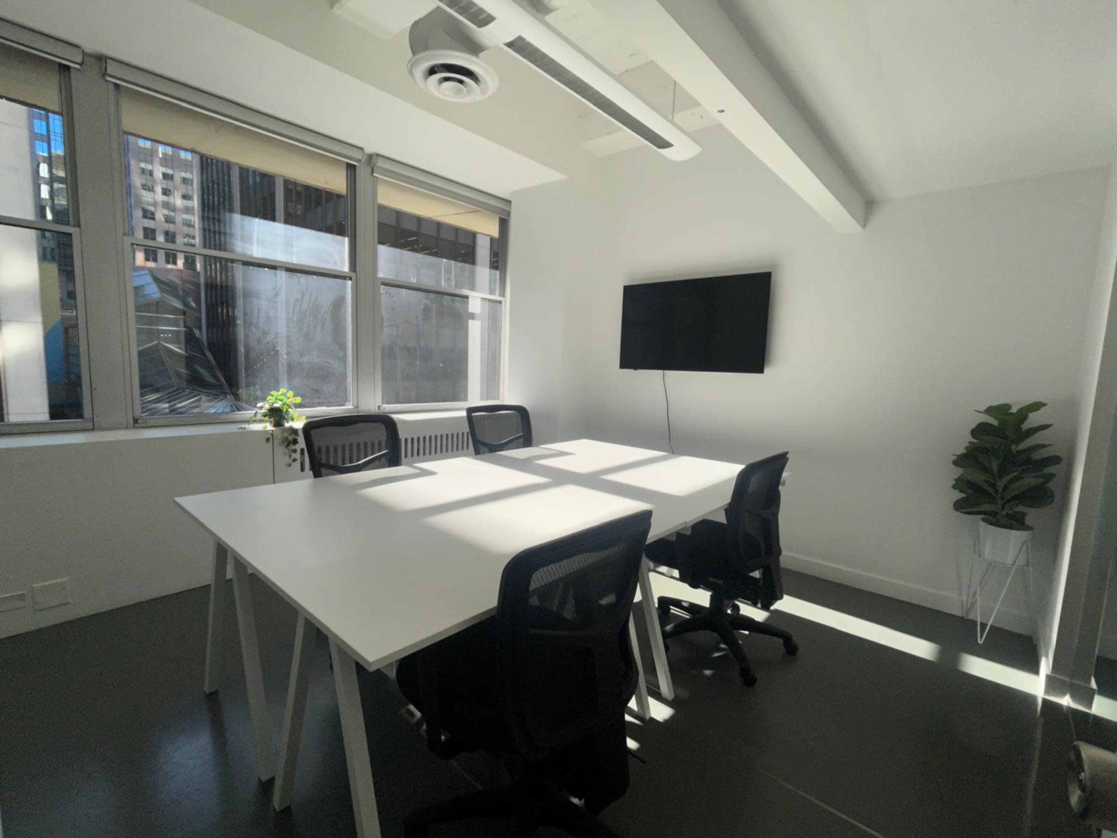 A modern conference room features a white table with four black chairs, large windows allowing natural light, and a wall-mounted television.