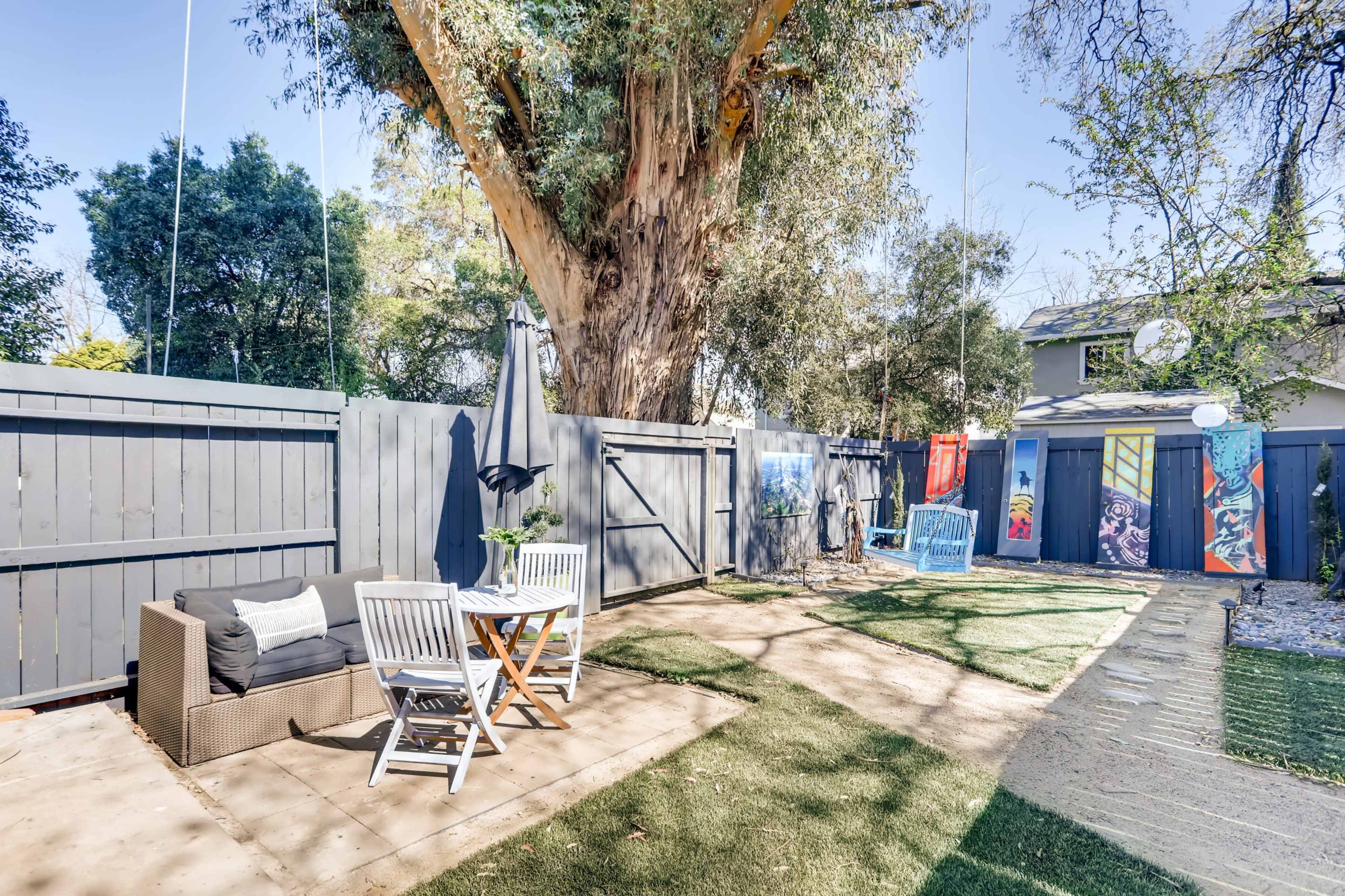 A patio area features a seating arrangement with a table and chairs, surrounded by greenery, under a large tree.