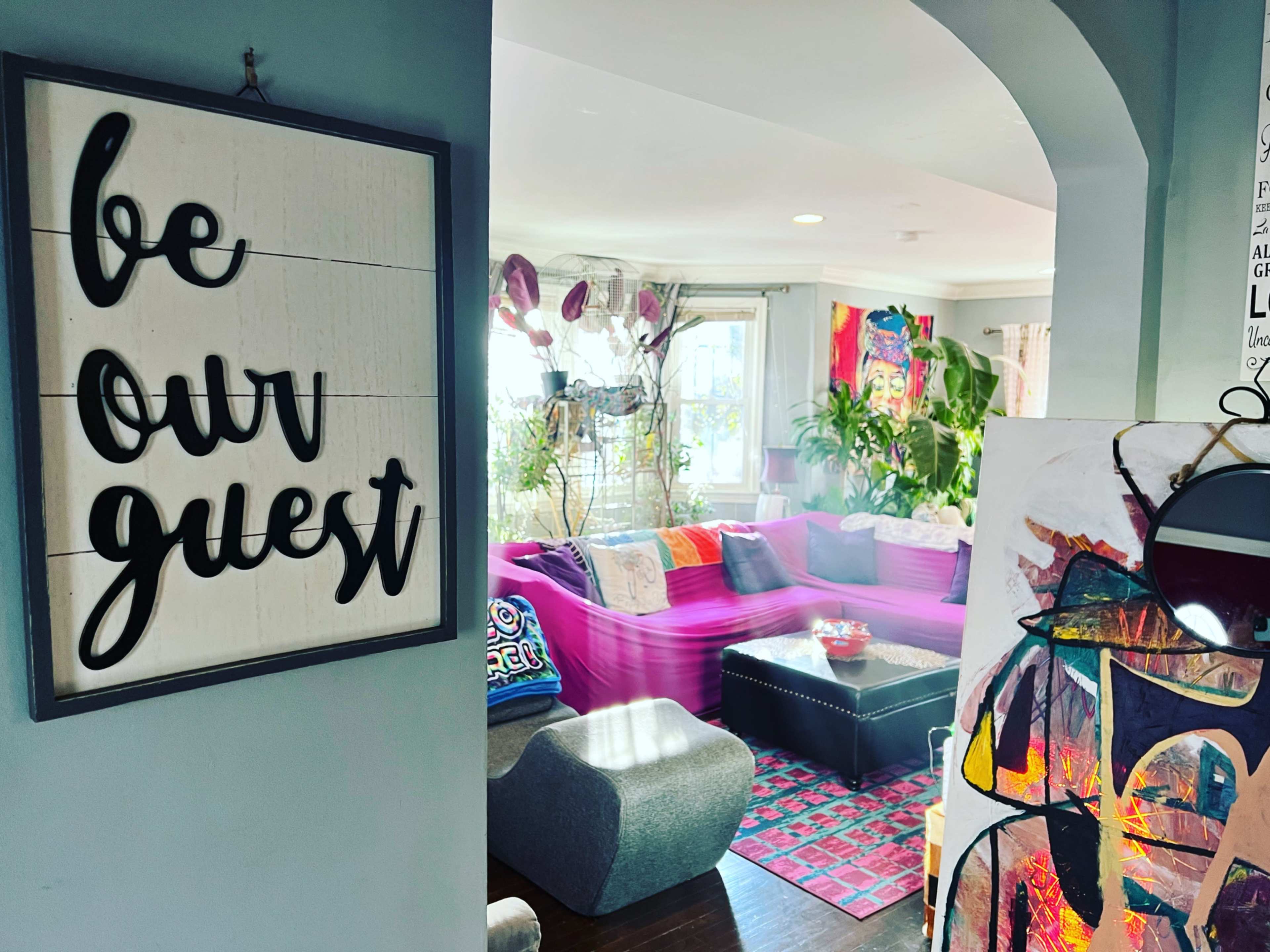 The image shows a welcoming living room with vibrant decor, plants, and a sign that reads "be our guest" on the wall.