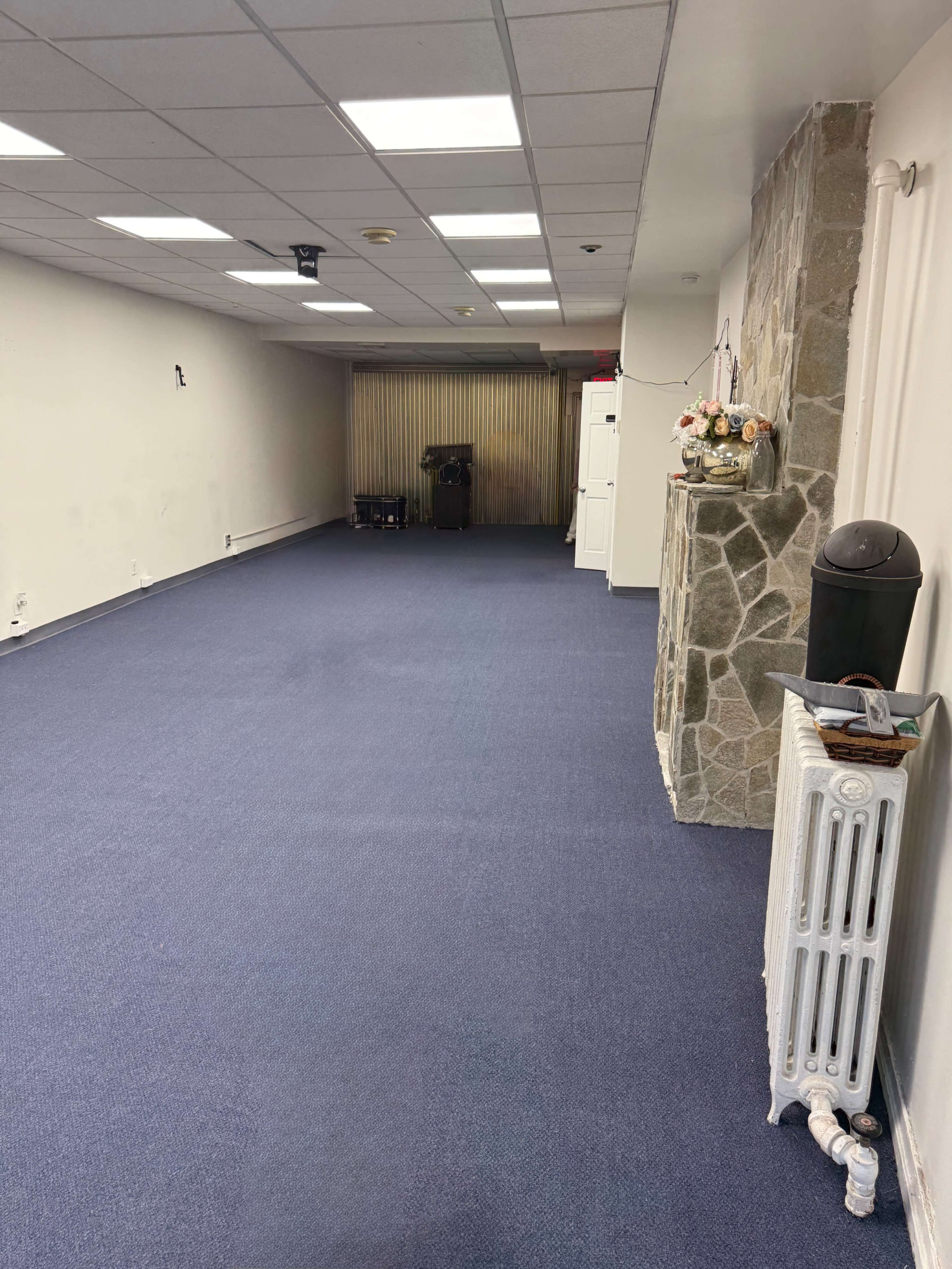 The image shows a spacious, empty room with blue carpeting, fluorescent ceiling lights, and a stone wall feature on one side.