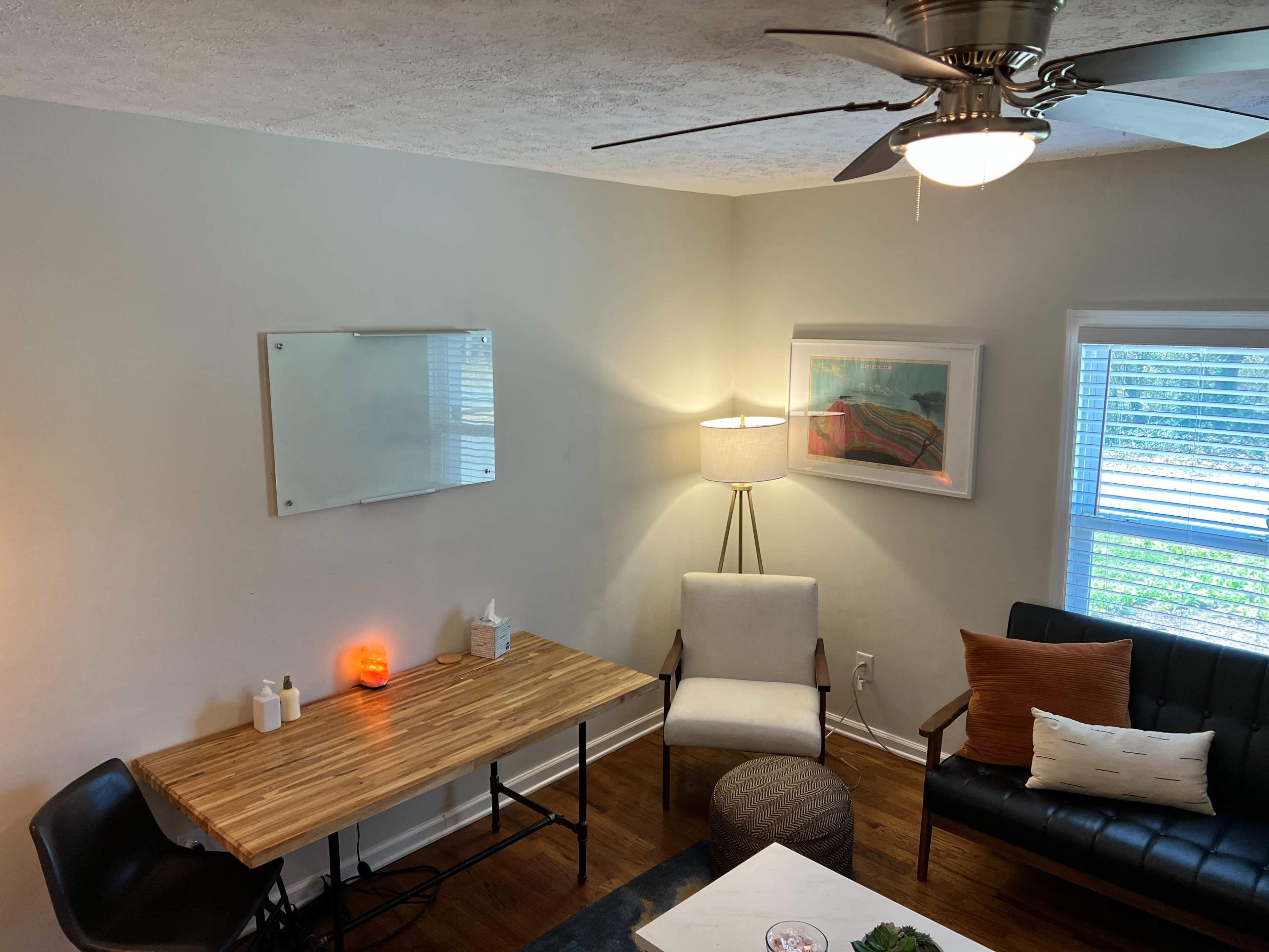 A minimalist living space features a wooden desk, an armchair, a black sofa, a lamp, and a wall-mounted whiteboard.