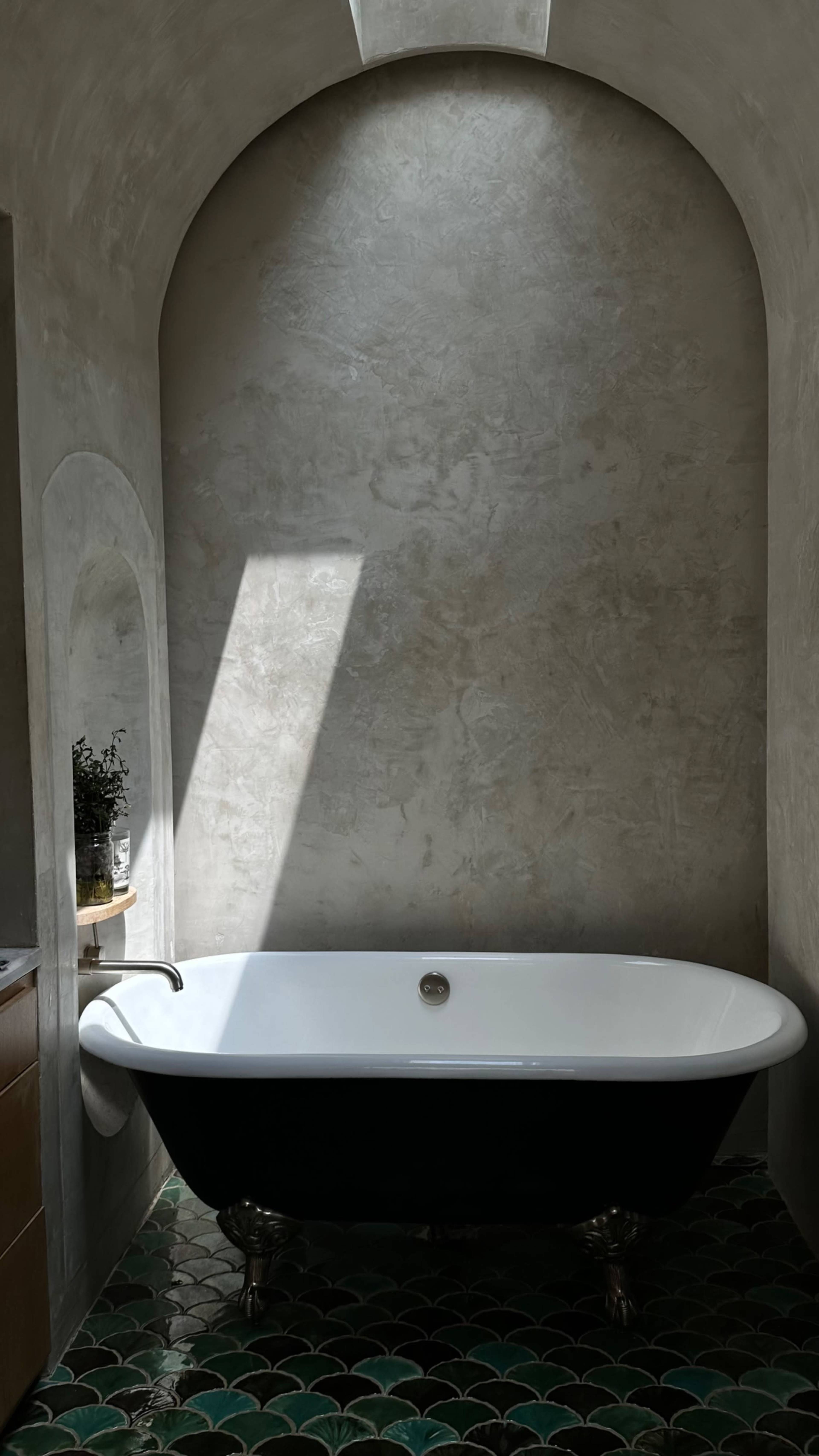 A freestanding bathtub is positioned under a beam of light in a minimalist bathroom with textured walls.