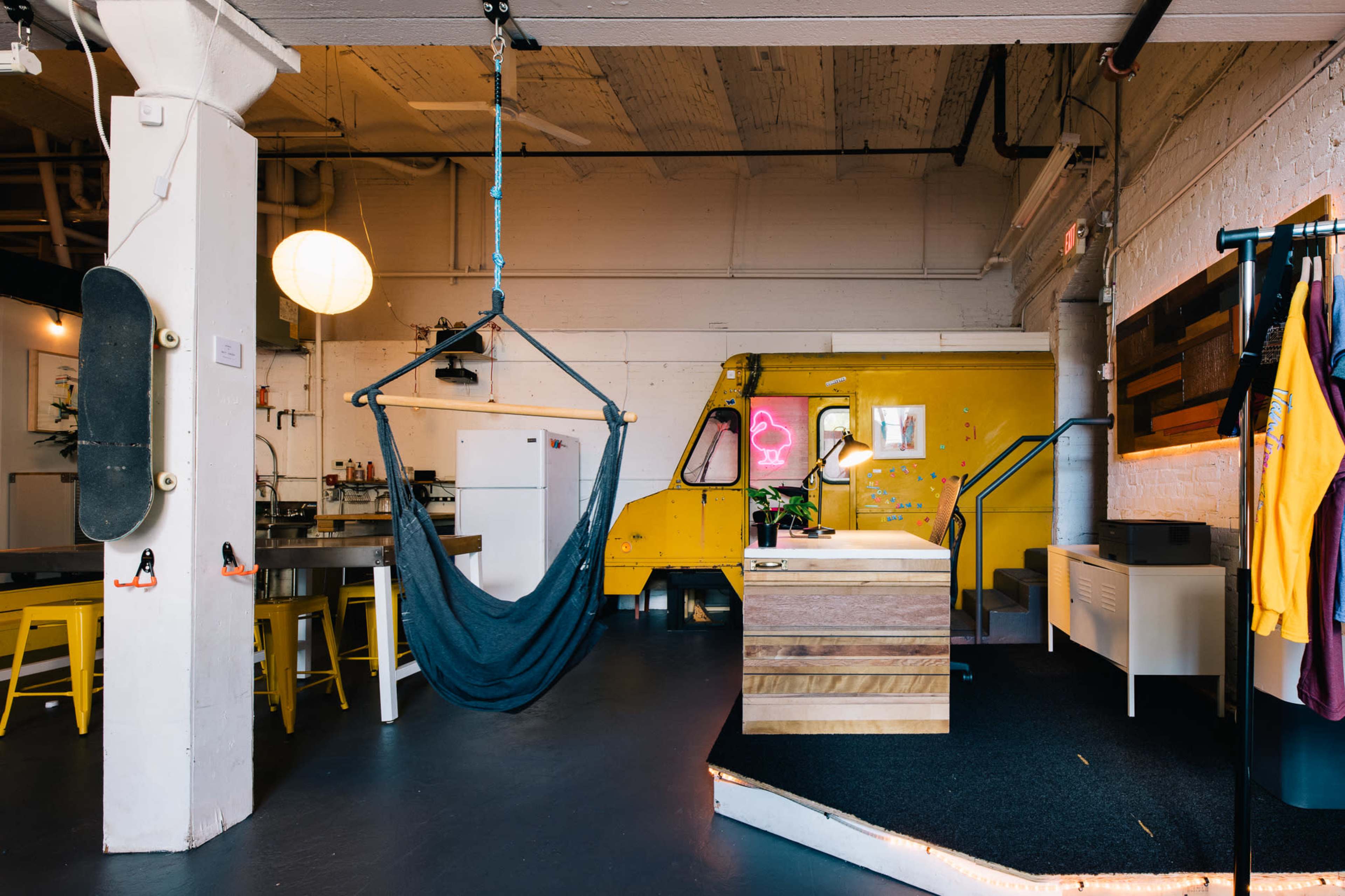 A modern interior space featuring a hammock suspended from the ceiling, a small kitchen area, and brightly colored decor including a yellow wall and neon lighting.