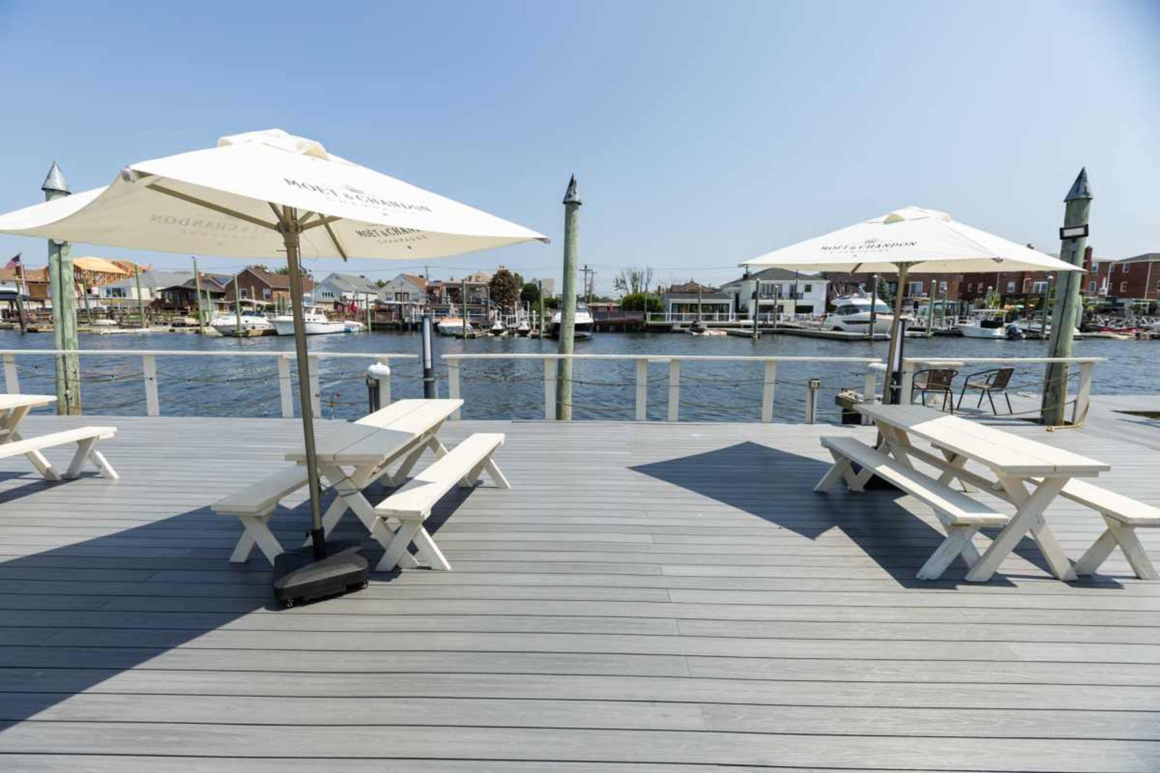 The image shows a waterfront deck with white picnic tables and umbrellas, overlooking a calm marina with boats.