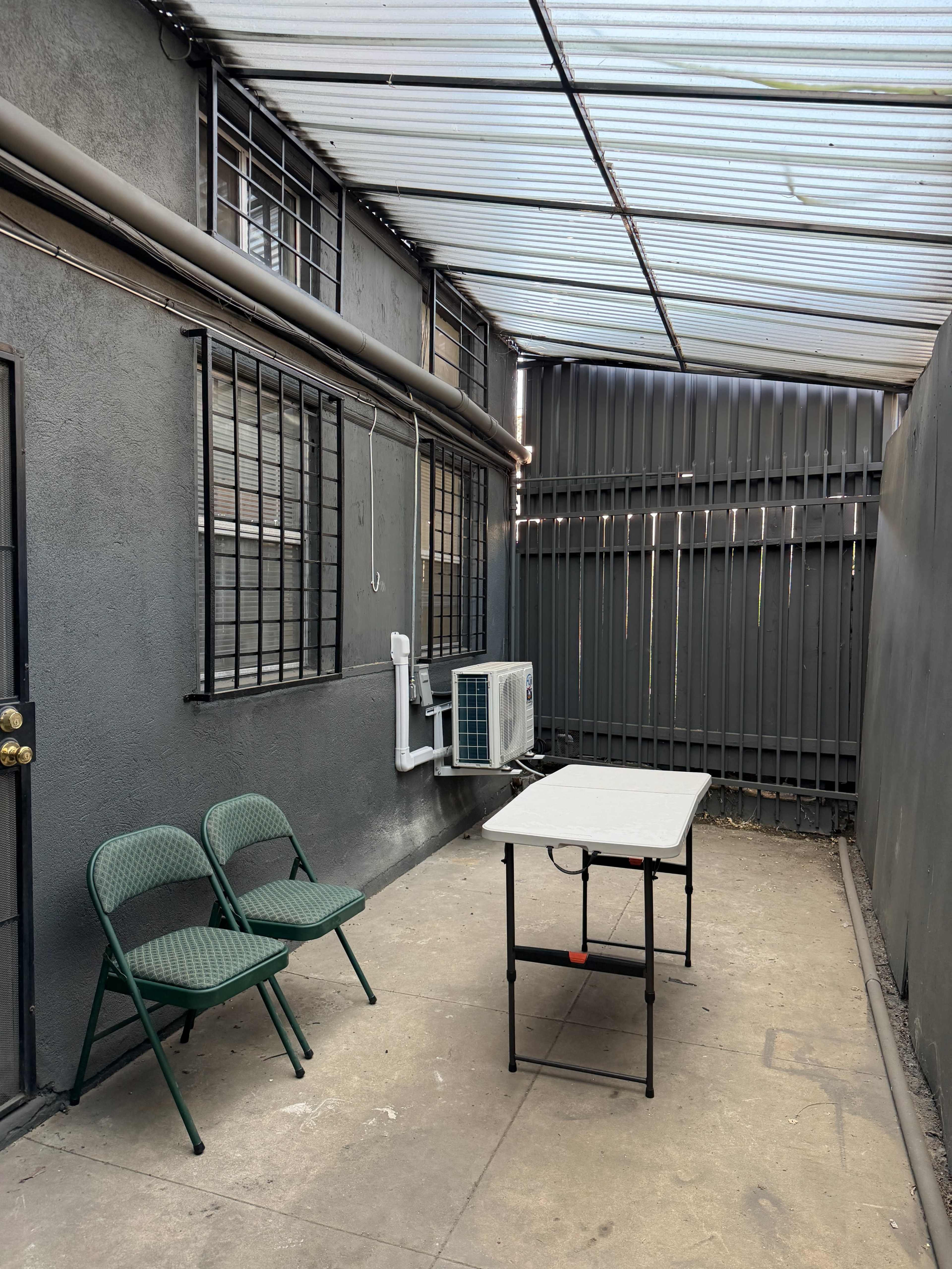 The image shows a small, enclosed patio featuring two green folding chairs and a folding table, with a wall-mounted air conditioning unit and metal fencing in the background.