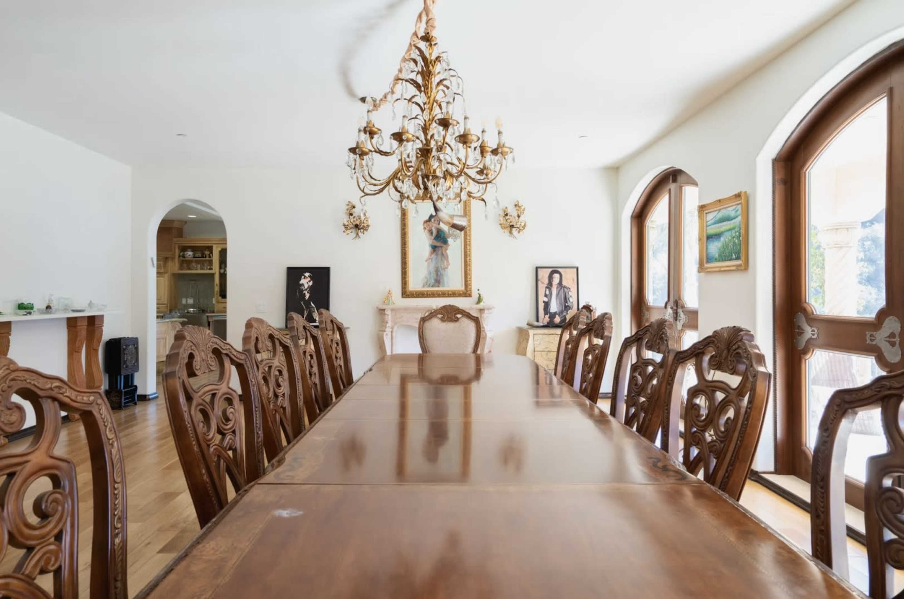 A long, polished wooden dining table is surrounded by intricately carved chairs beneath a chandelier in a well-lit dining room featuring arched windows and artwork on the walls.