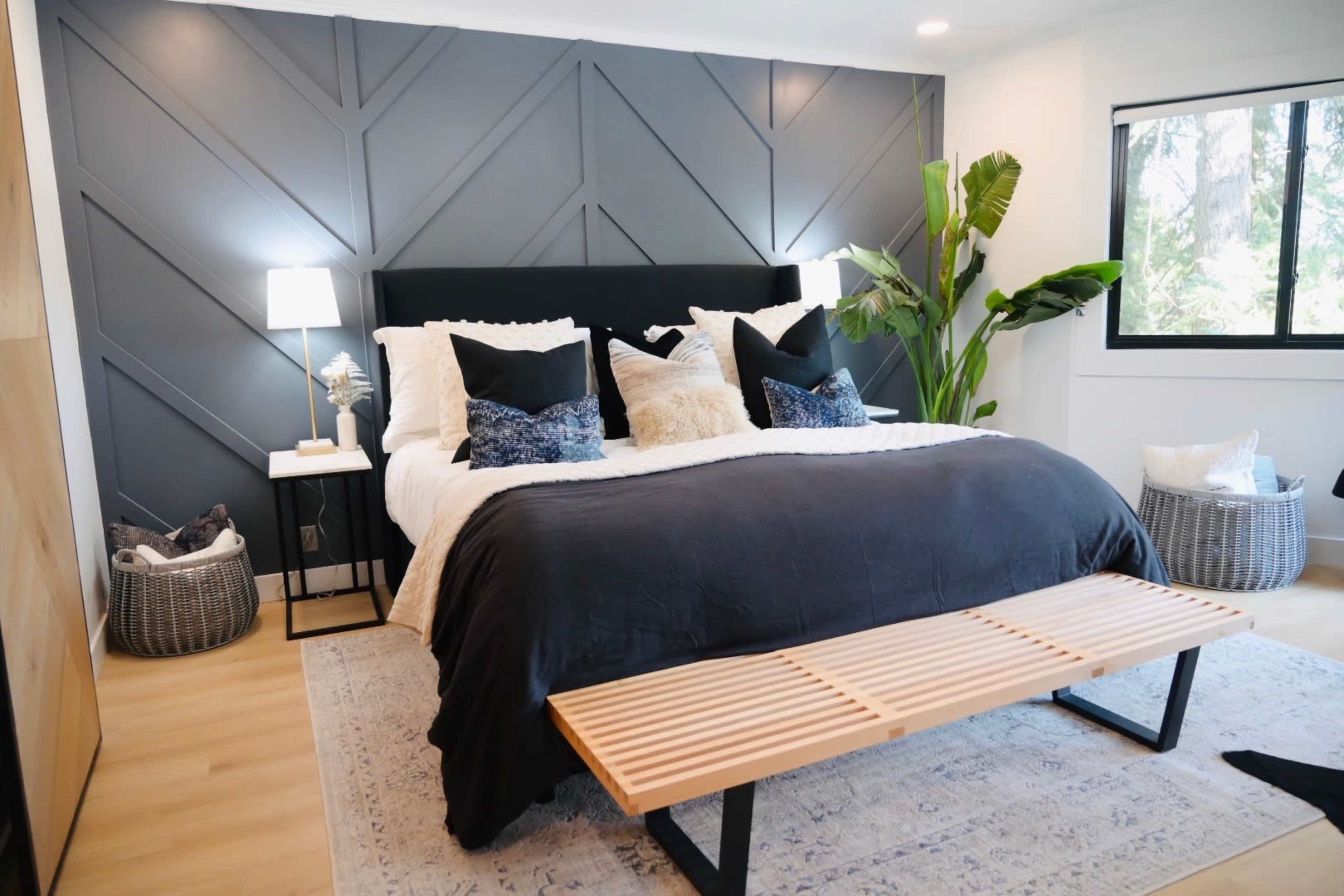 The image shows a modern bedroom featuring a large bed with dark bedding, decorative pillows, a wooden bench at the foot, and a wall with a geometric design.