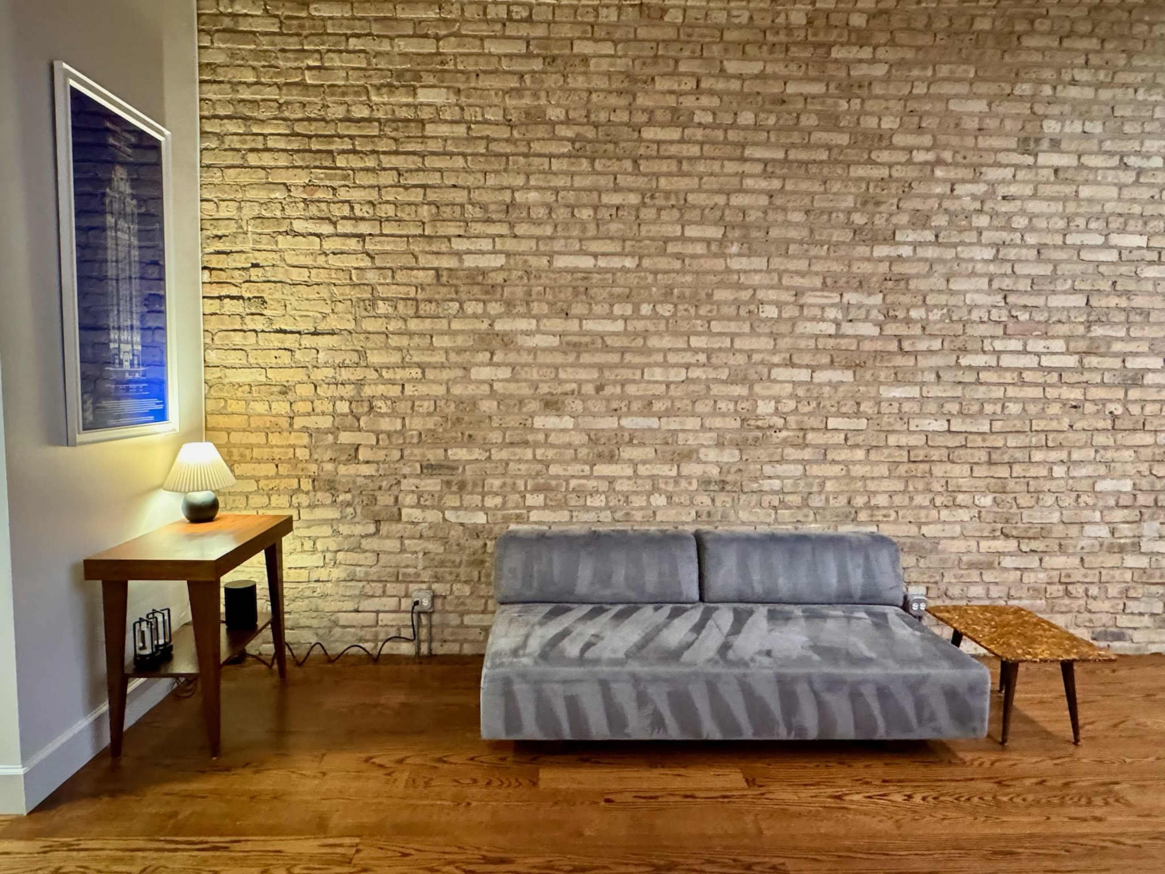 A gray sofa sits against a exposed brick wall, accompanied by a small wooden table and a lamp.