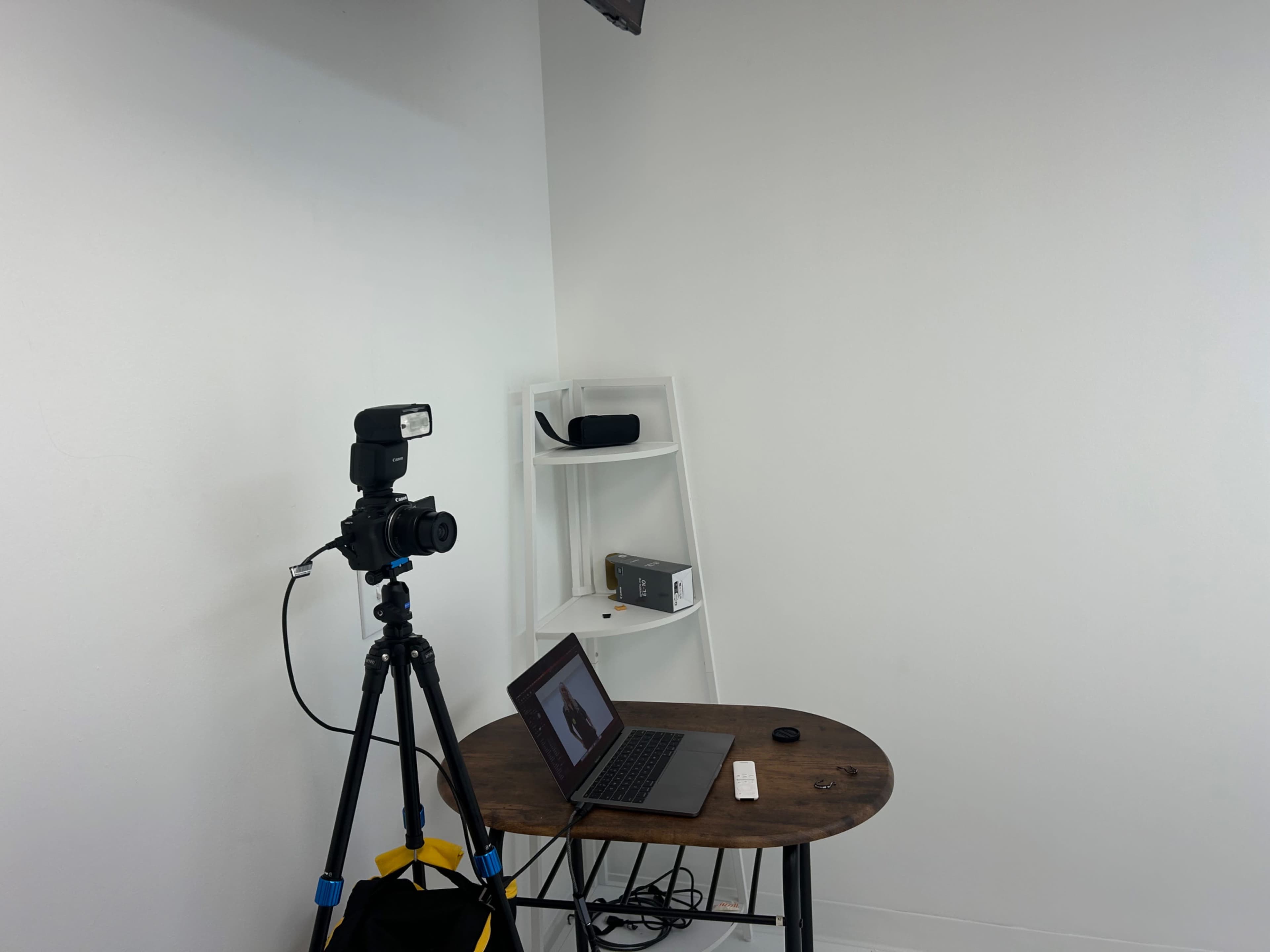 A camera on a tripod is set up next to a small table with a laptop and accessories against a white wall.
