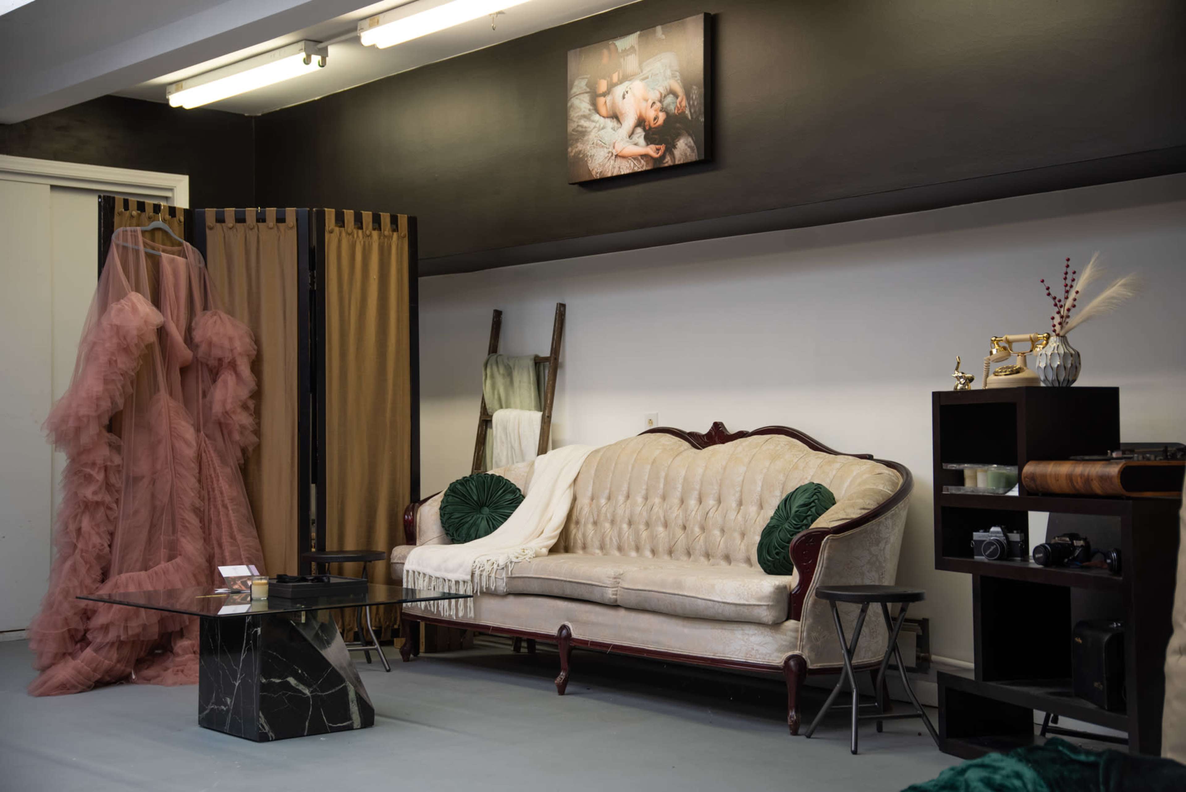 A stylish room featuring a vintage couch, a large pink dress on a mannequin, a black side table, and various decorative items on shelves.