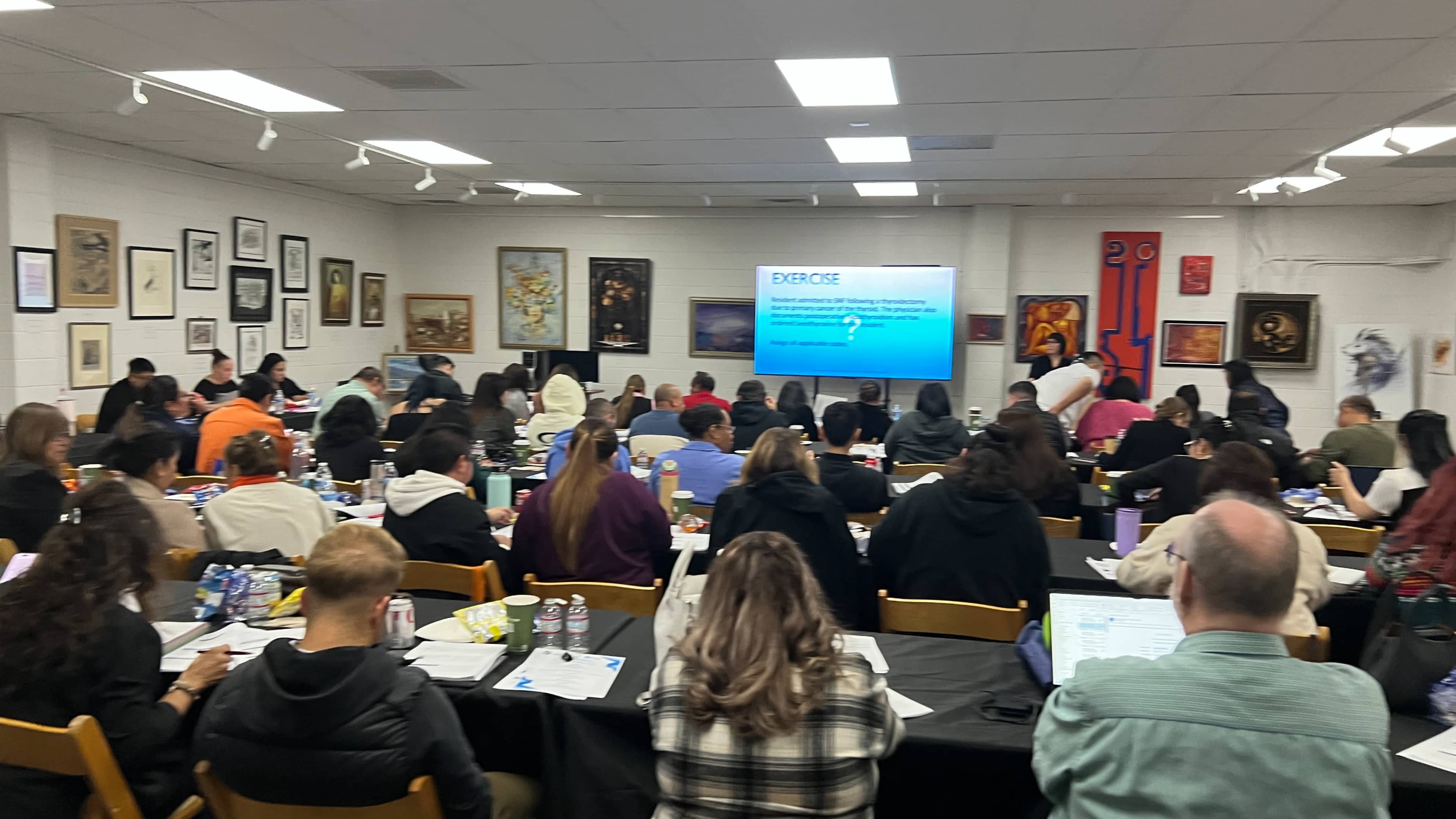 The image shows a classroom filled with students seated at tables, engaged in a presentation displayed on a screen at the front.