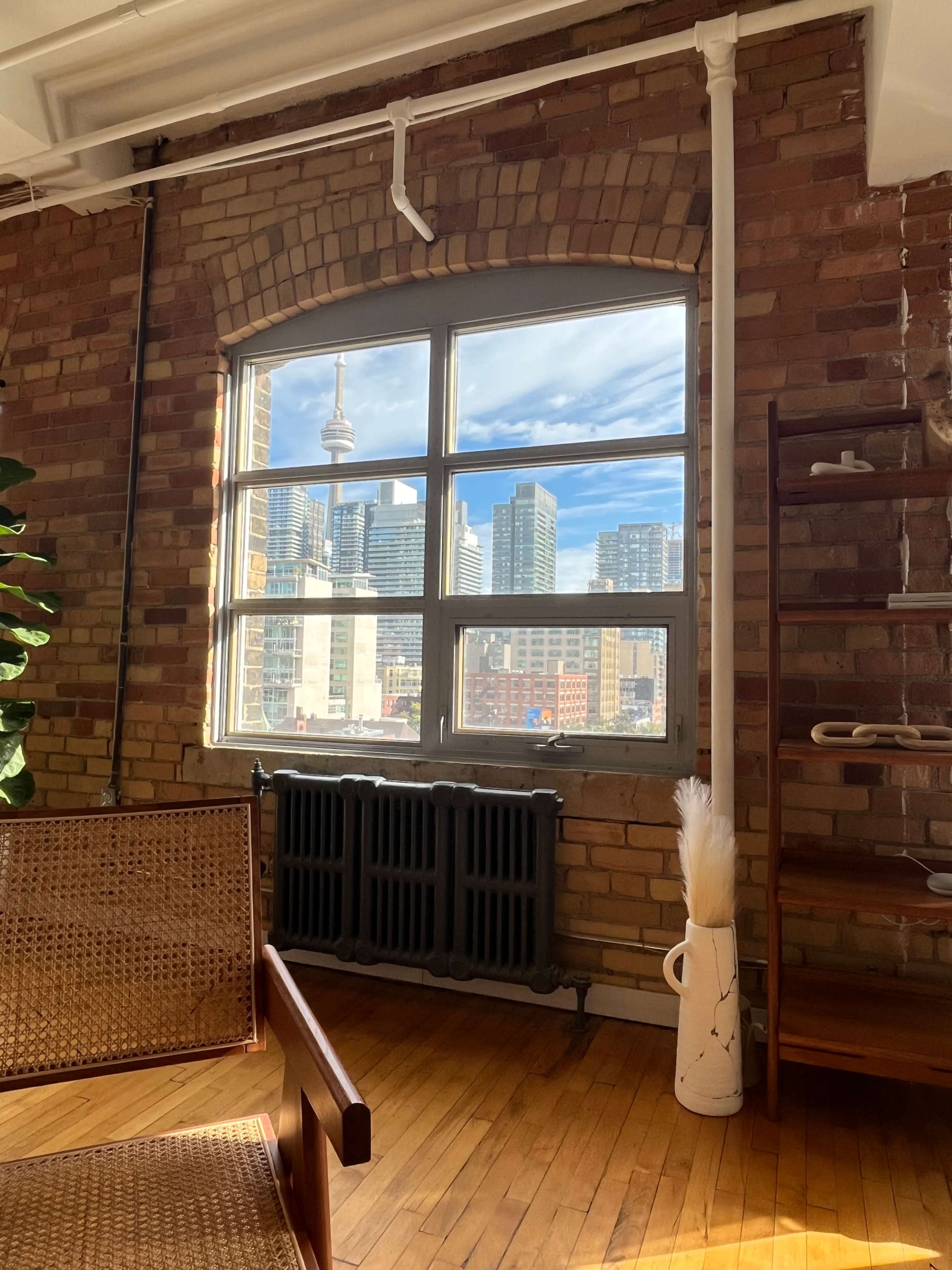 A view from a room featuring a large window that overlooks a city skyline with tall buildings, including a prominent tower, framed by a rustic brick wall.