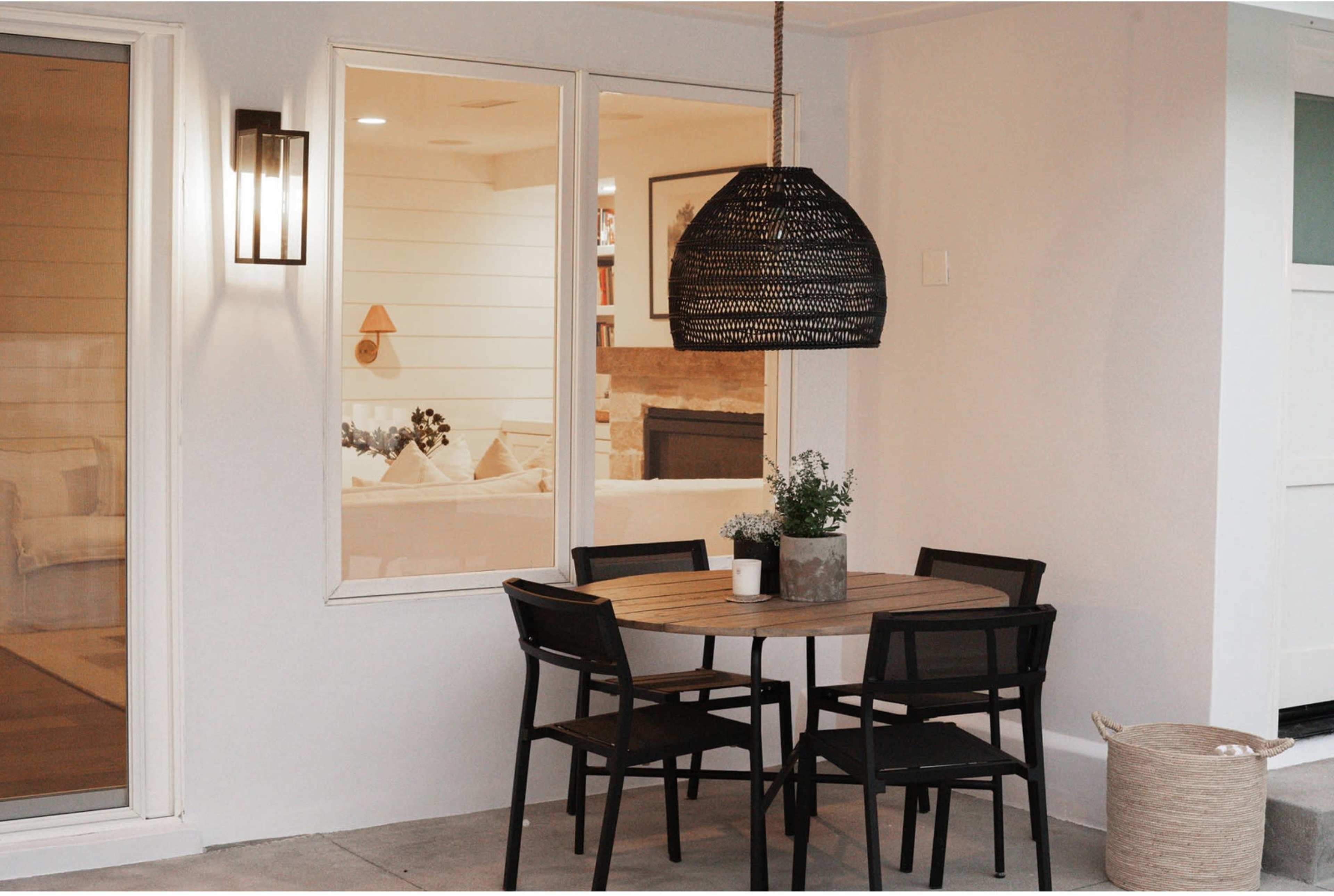A small outdoor dining area features a round wooden table with four black chairs, a hanging pendant light, and a potted plant, adjacent to a window displaying an interior living space.